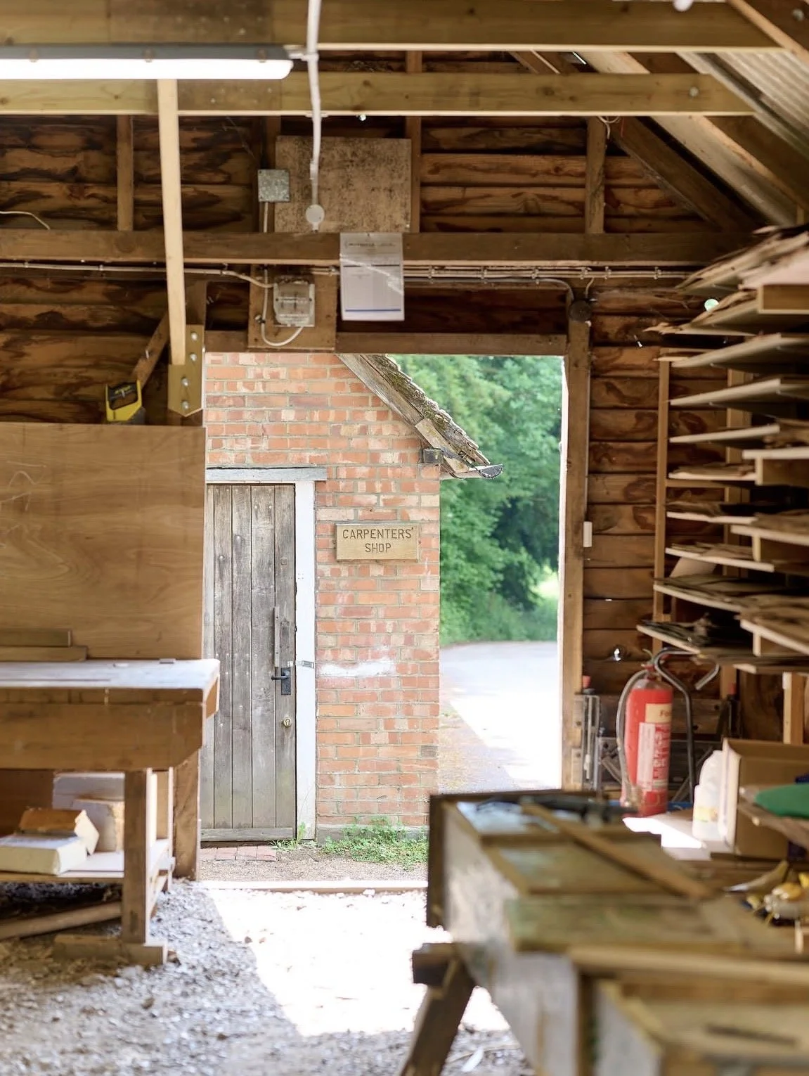Inside a woodworking shed with shelves filled with wood pieces, a worktable, and a fire extinguisher. Outside the open door, a brick building with a sign that reads 'Carpenters' Shop' is visible, along with a gravel pathway and green foliage.