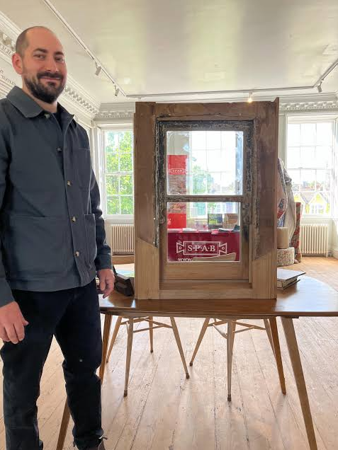 A man standing next to a small wooden structure on a table inside a bright room with large windows and wooden floors.