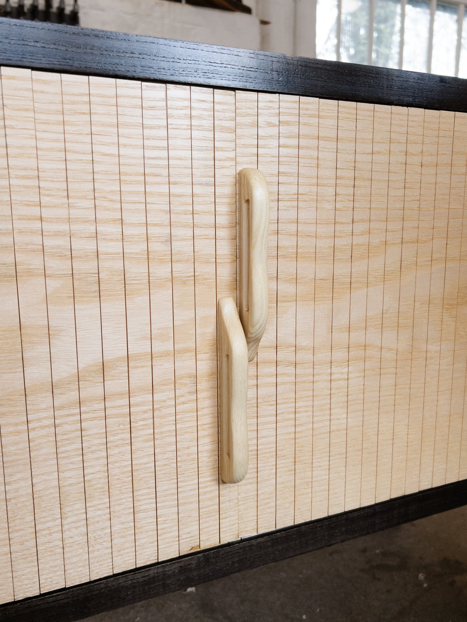 Close-up of a wooden cabinet with a black frame and a wooden handle on the door, placed on a dark surface with a window and trees visible in the background.