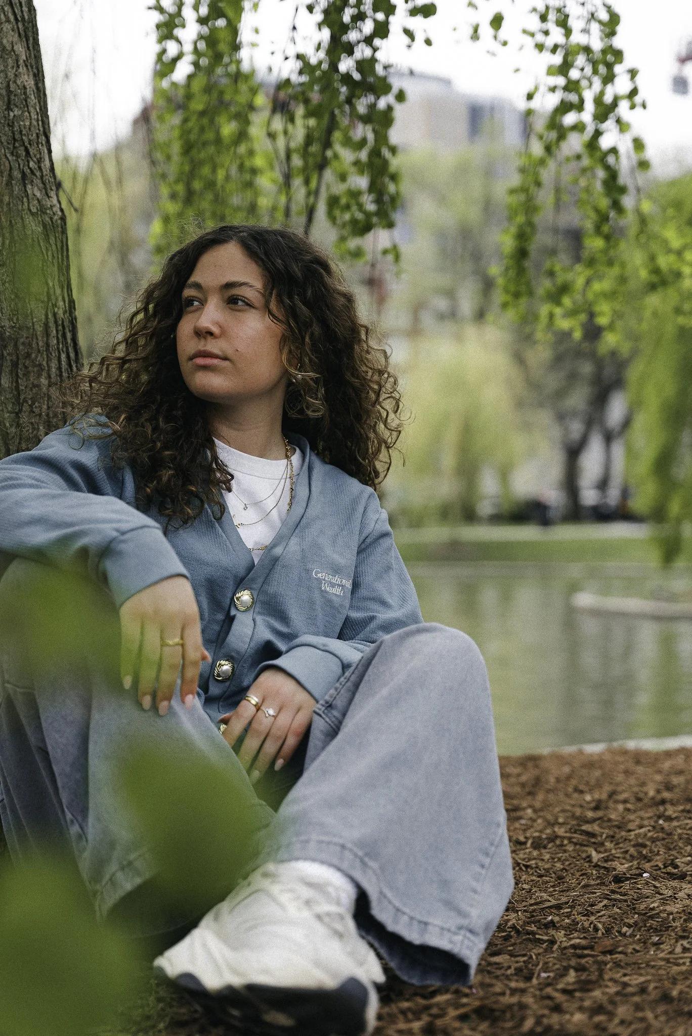 A young woman with curly hair sitting by a tree on a park bench, wearing a blue jacket and gray pants, with a body of water and green trees in the background.