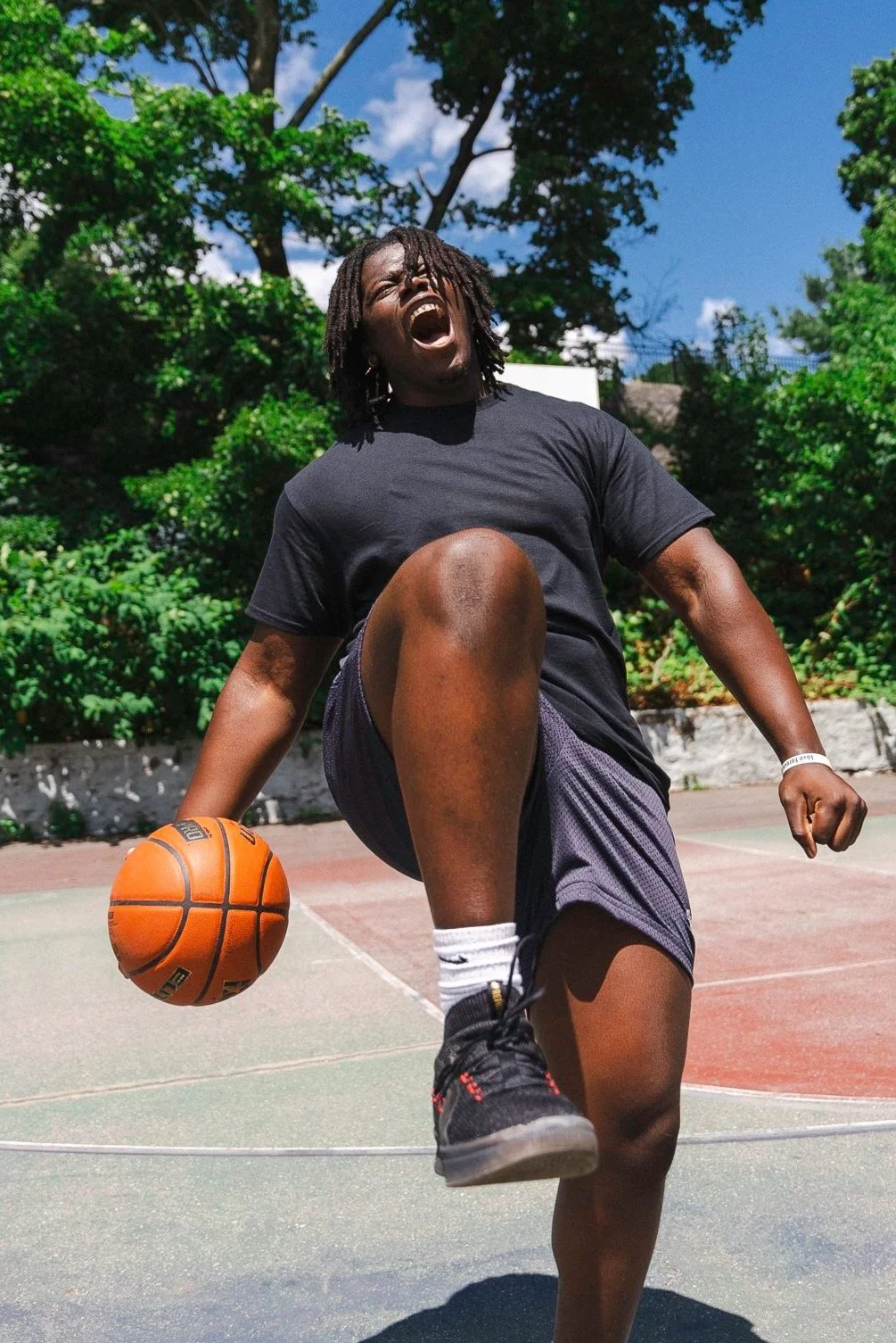 A young man playing basketball outdoors on a court, mid-move with his mouth open, wearing a black T-shirt, shorts, and black sneakers, holding a basketball, with trees and a blue sky in the background.