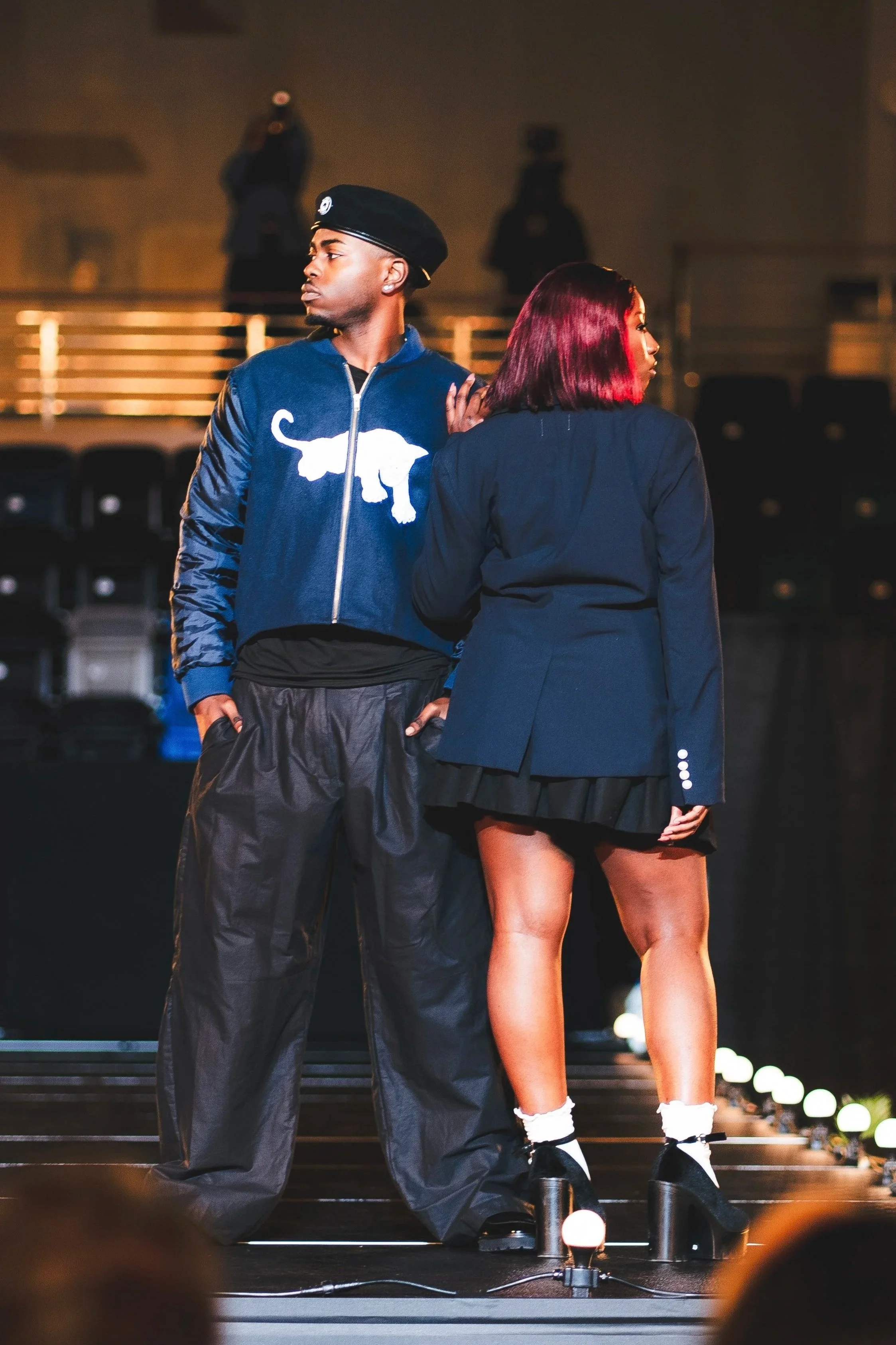 Two young adults on stage, facing away from each other, one wearing a blue Puma jacket and black pants, the other wearing a navy blazer and a black skirt, with stage lights and audience in the background.