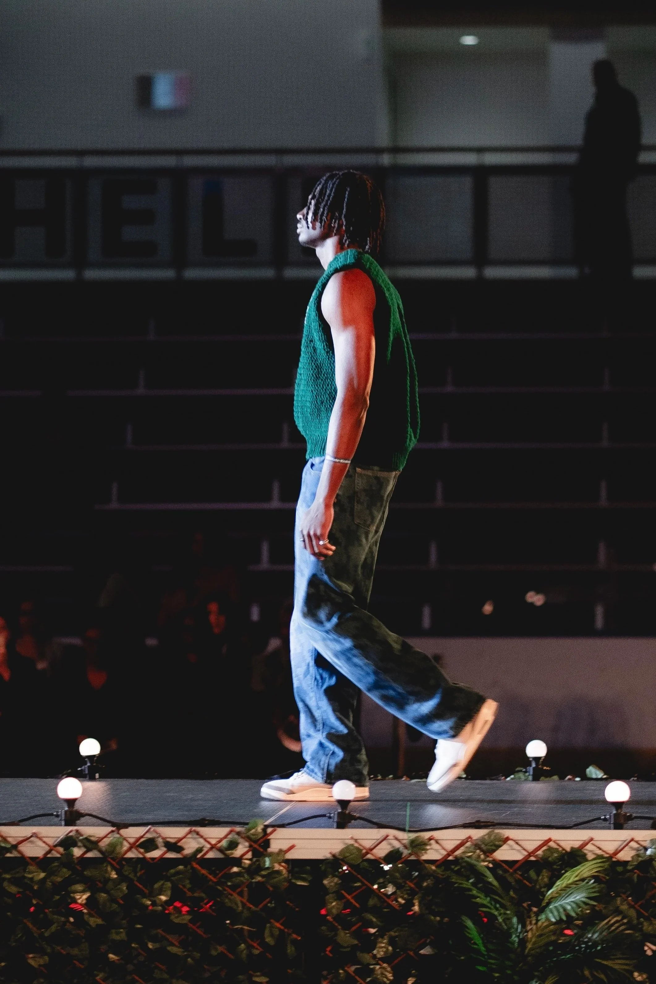 A person with dreadlocks walking on a runway during a fashion show, wearing a green sleeveless sweater, baggy jeans, and white sneakers under stage lights.