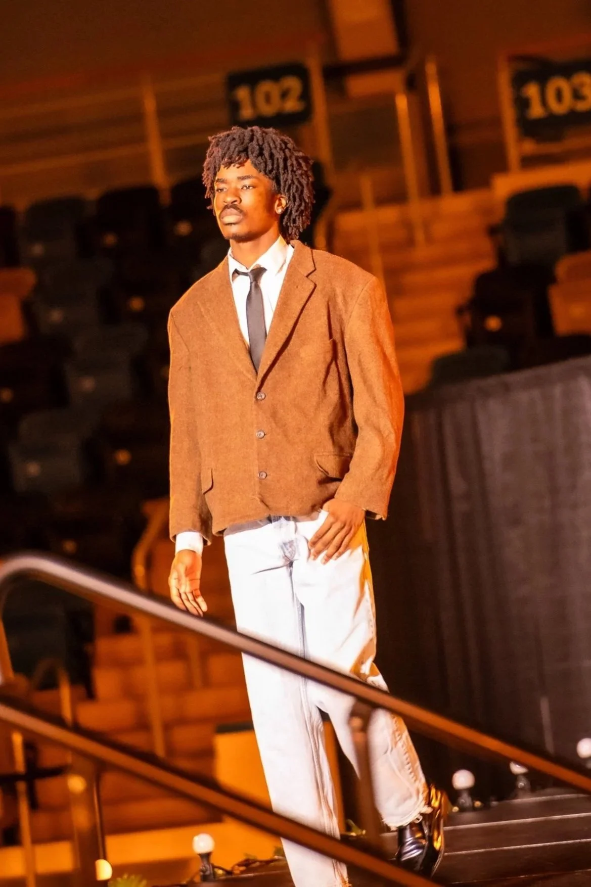 A man with curly hair wearing a brown jacket, white shirt, black tie, and white pants standing on an indoor stage or auditorium with wooden steps and black chairs in the background.