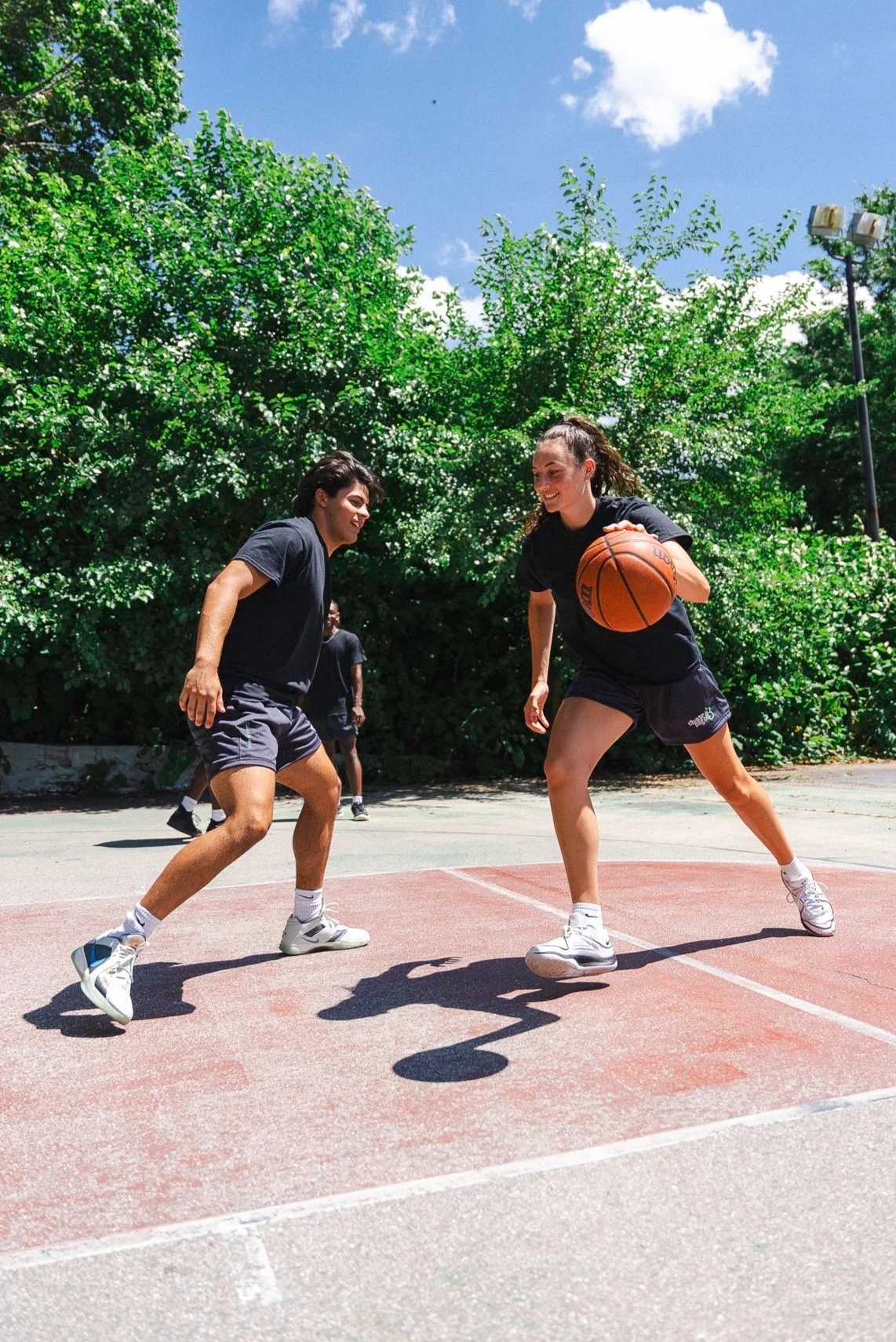 Two young women playing basketball on an outdoor court, with greenery and blue sky in the background.