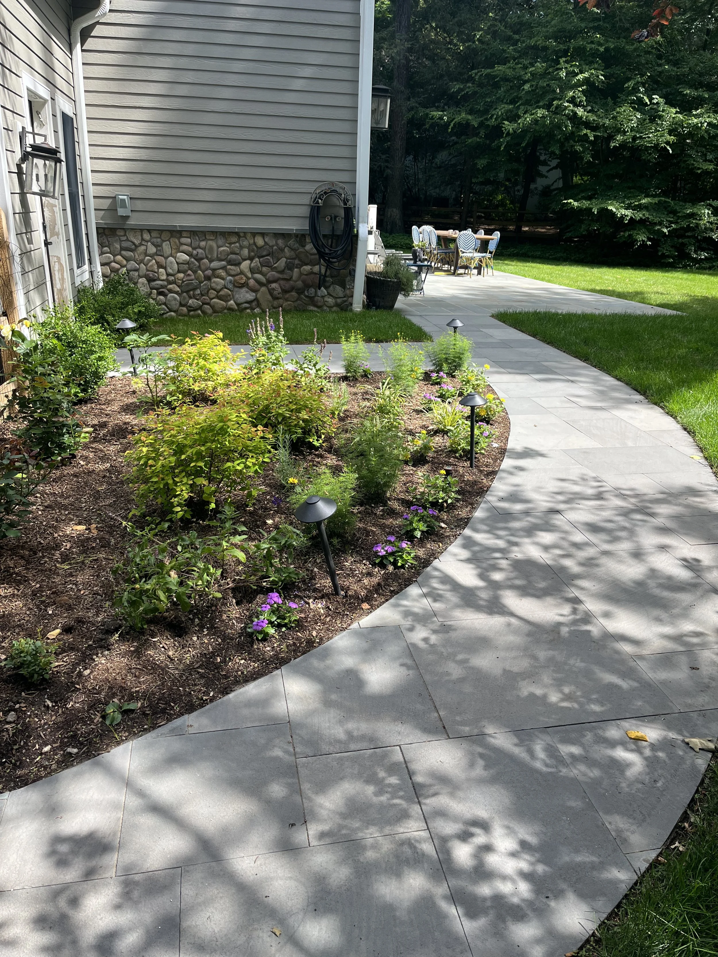 A backyard patio area with a curved concrete path, garden bed with various plants and flowers, outdoor table with chairs, and a house with siding and stone foundation. Trees and greenery are in the background. in Franklin lakes NJ 