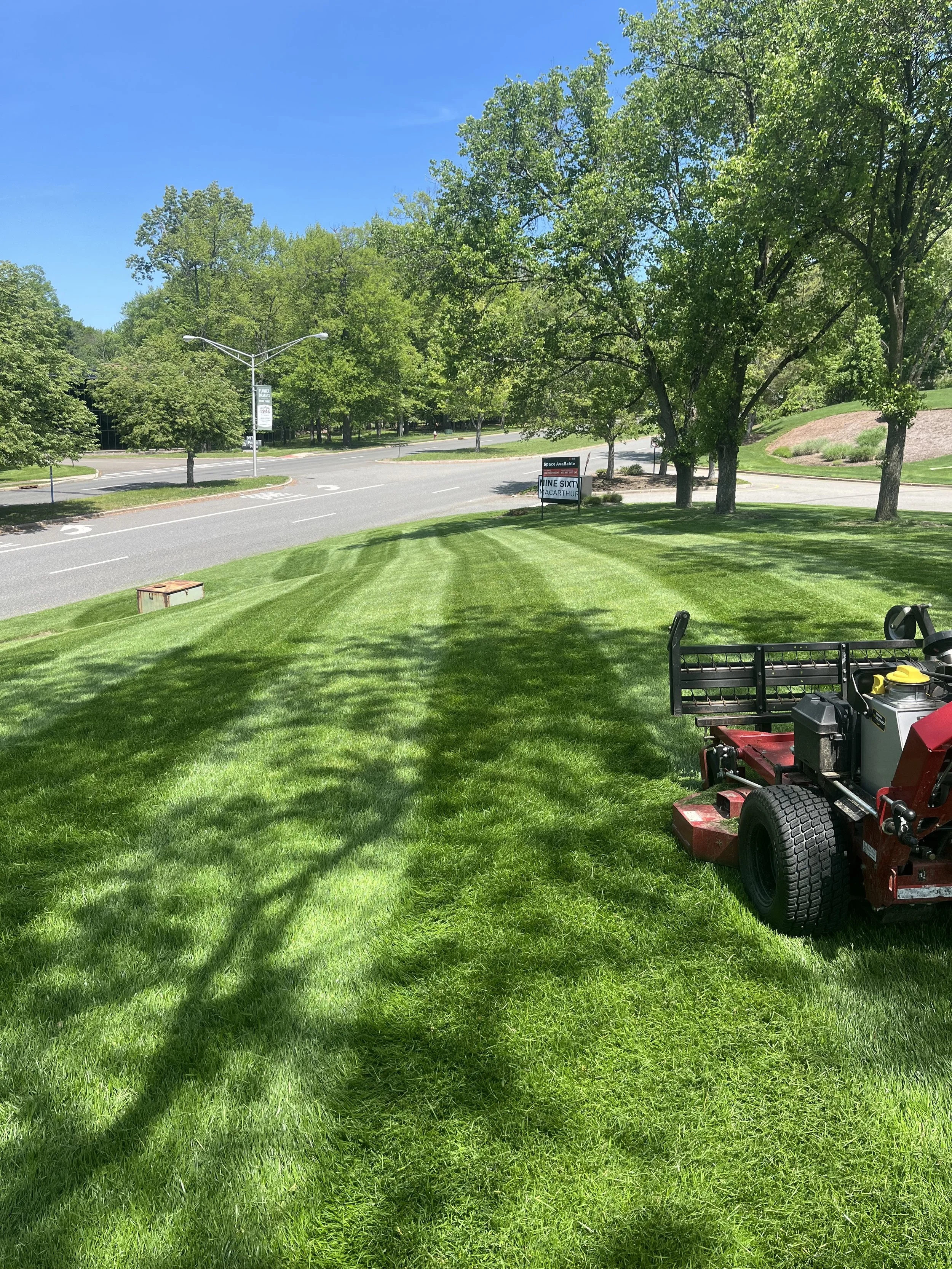 A well-maintained green lawn with a push lawn mower on it, trees providing shade, a street with cars and street signs in the background, and a sign advertising space for lease.