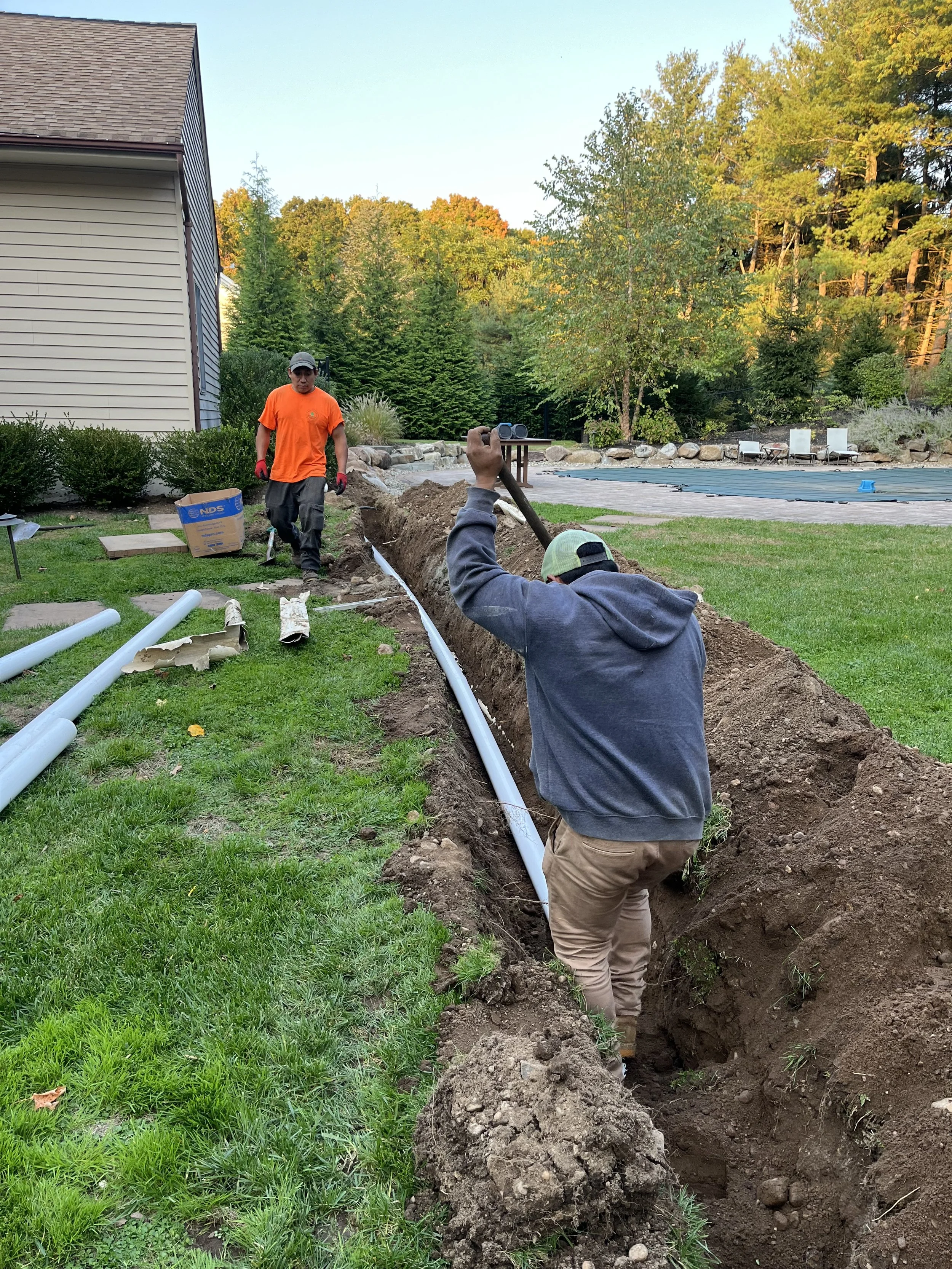 Two men working on installing underground pipes in a backyard, with a worker holding a shovel and another digging in a trench, surrounded by grass, trees, and backyard furniture.