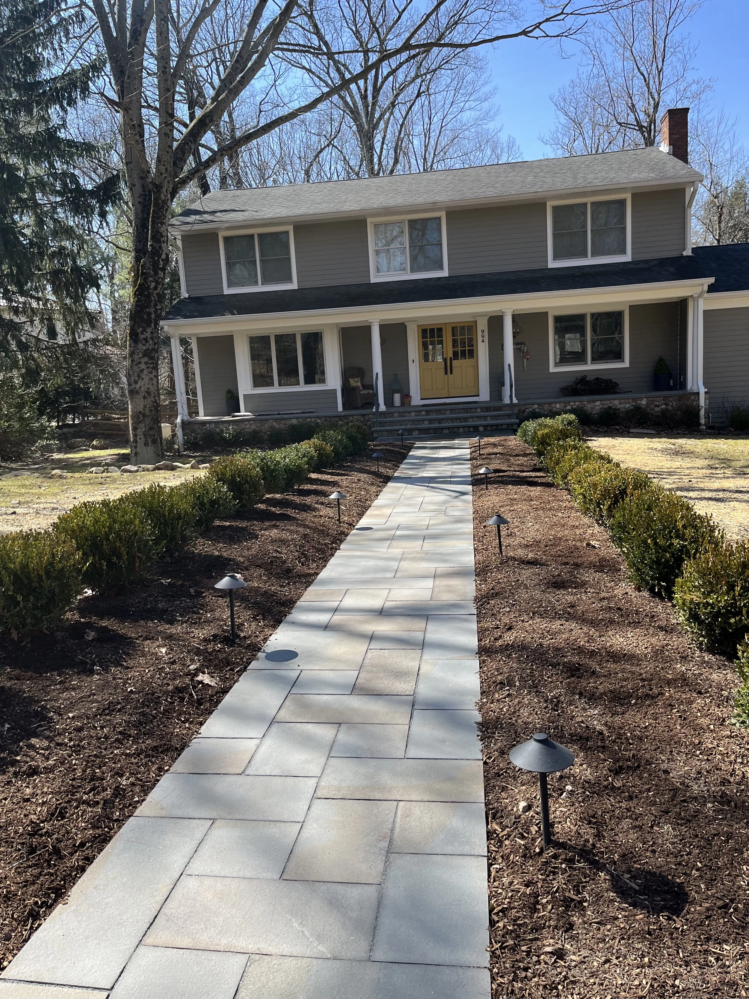 Newly paved walkway leading up to a house entrance, with small landscape lights on each side, a lawn, and a large tree in the front yard. The house has grey siding, white trim, a yellow front door, and a porch with white columns. in Franklin lakes nj