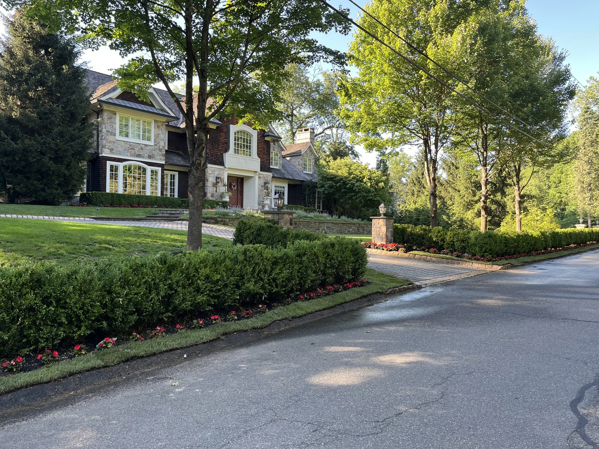 A large residential house with stone and dark wood exterior, surrounded by trees and a well-maintained front yard with grass, shrubs, flowers, and a brick walkway.