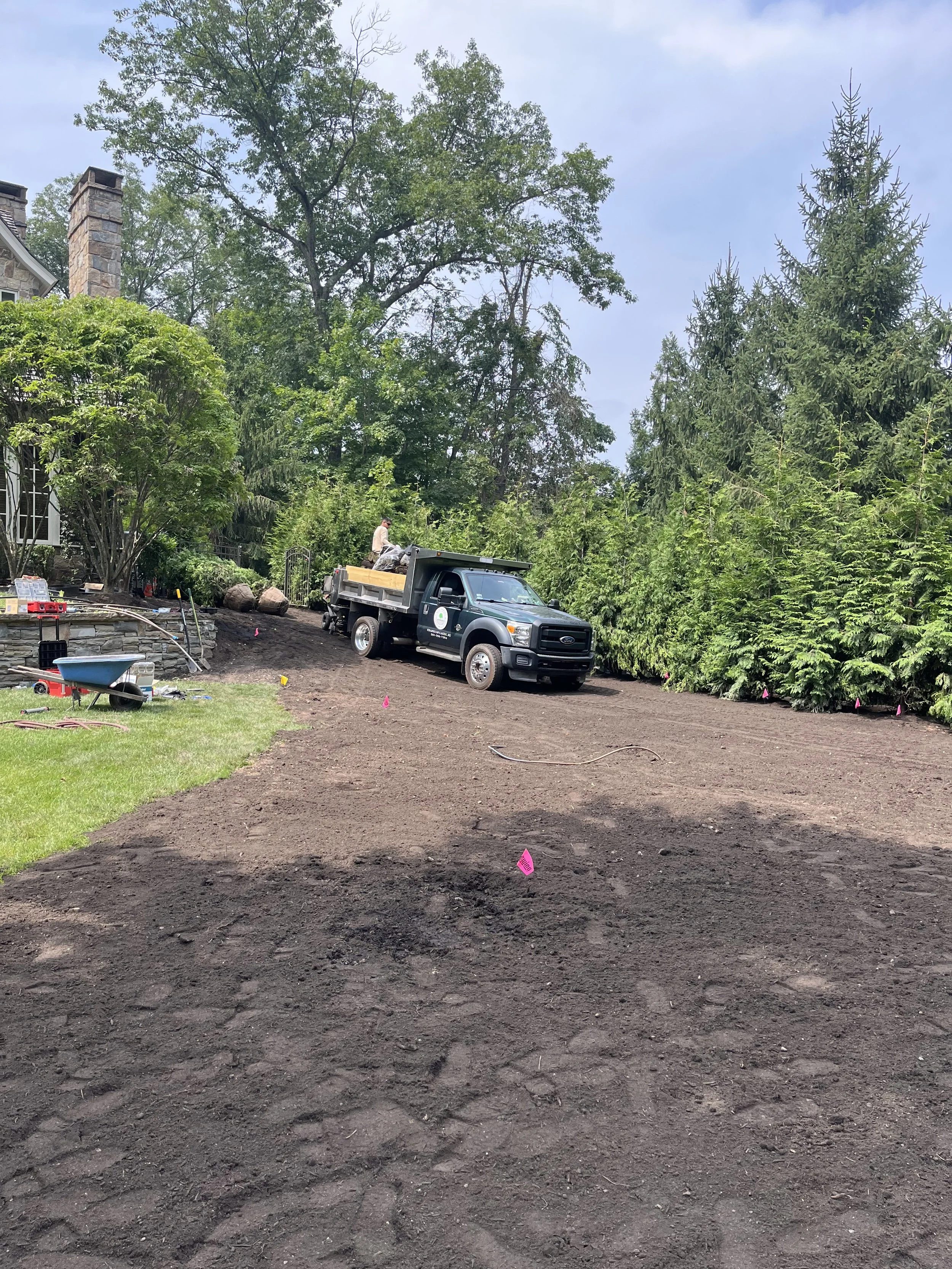A house with a stone chimney is visible on the left side behind trees. A black dump truck with a person on the bed is parked on a dirt driveway, which is marked with pink flag markers. There are several small trees or shrubs along the edge of the dri
