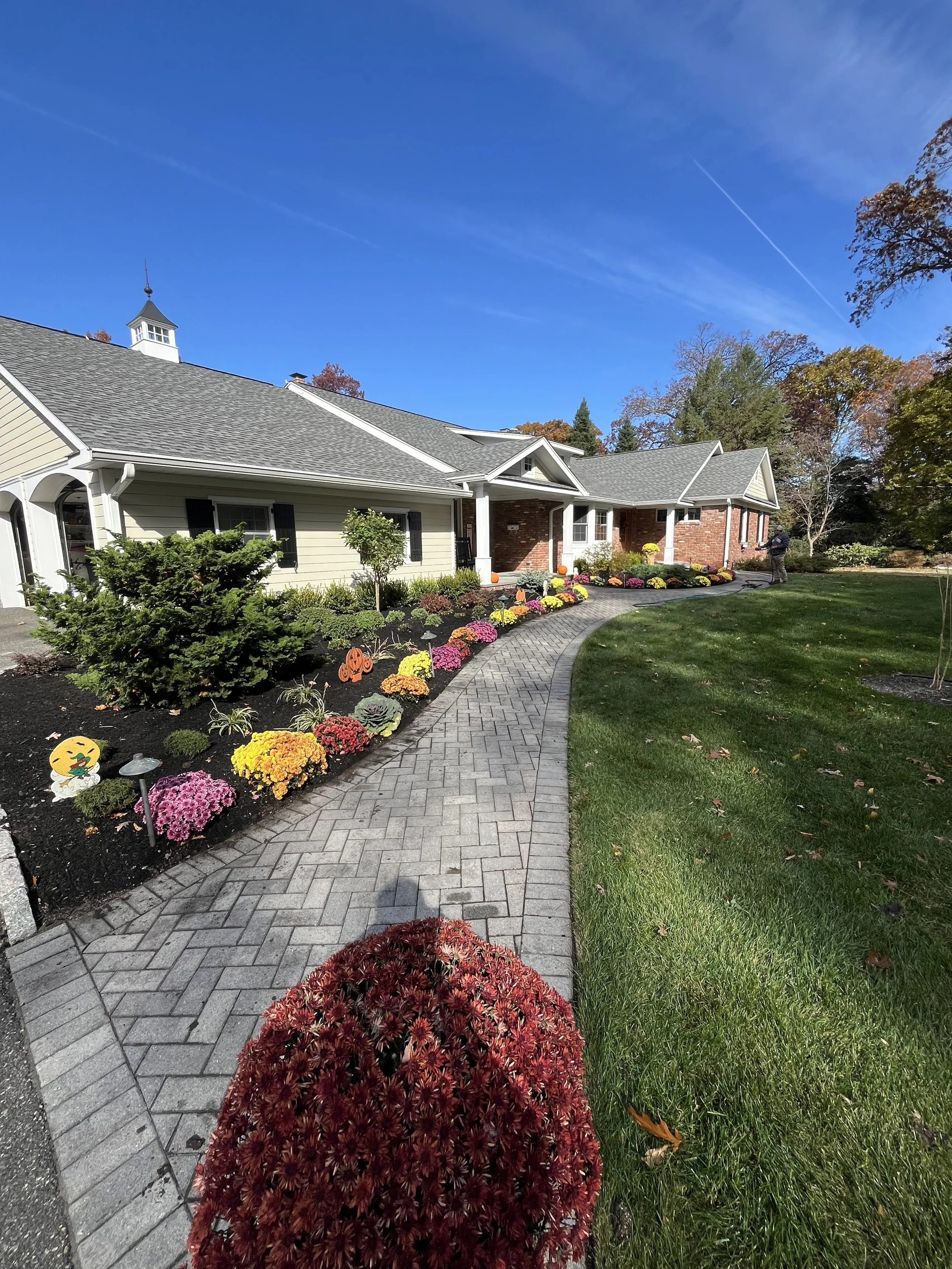 A house with a brick and siding exterior surrounded by a landscaped yard with colorful fall flowers and plants, a brick driveway, and trees with changing fall foliage under a clear blue sky. in wyckoff nj