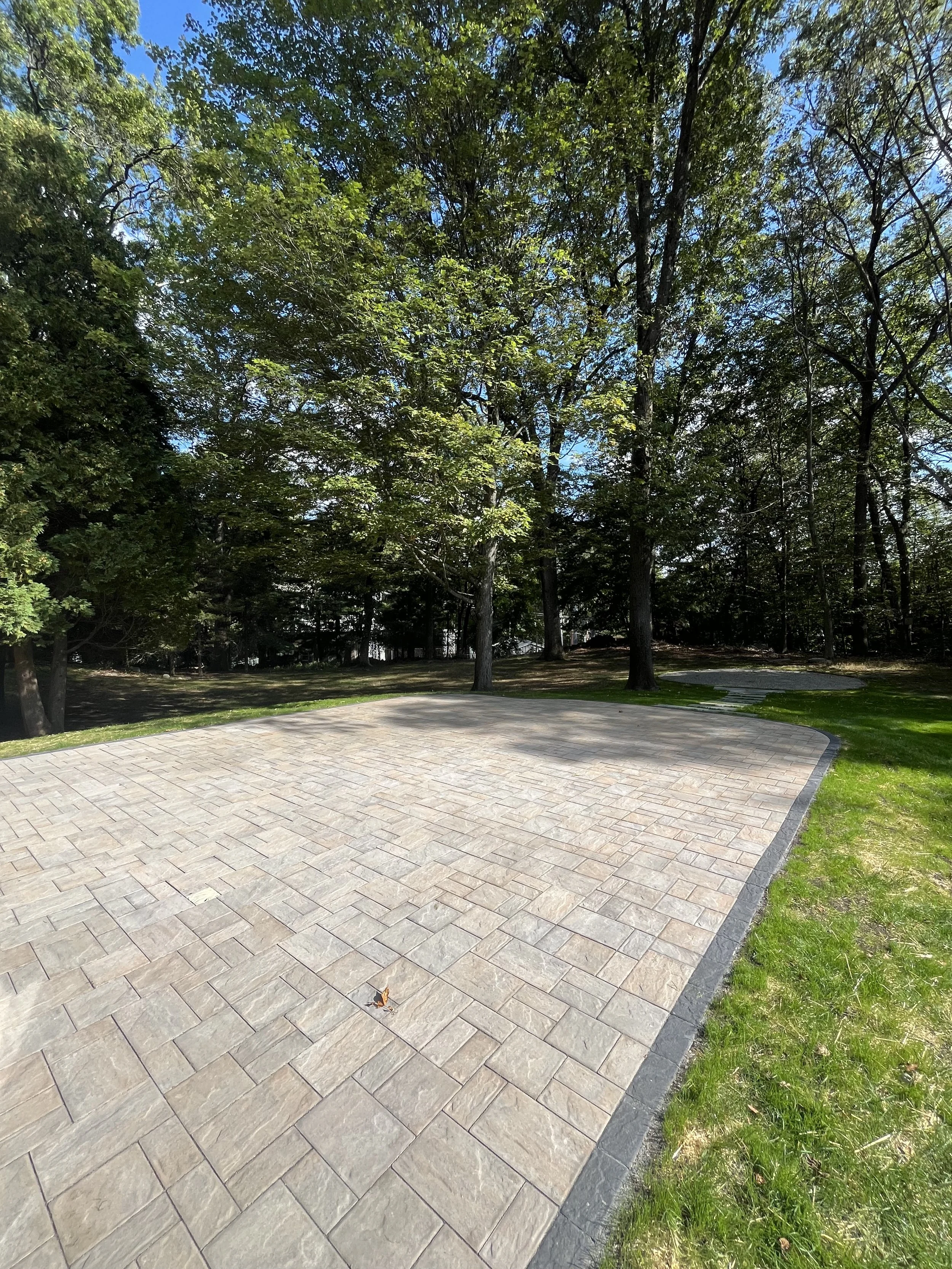 A paved stone pathway curves through a grassy area with tall trees with green leaves and a bright blue sky overhead.