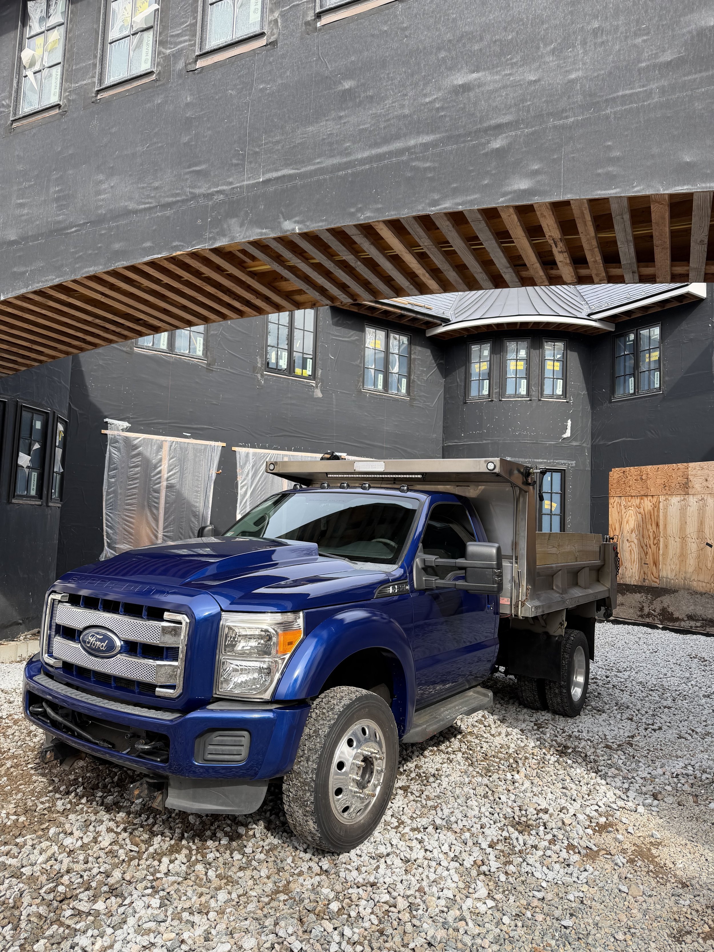 A blue Ford pickup truck parked on gravel in front of a building under construction, with black exterior walls and multiple windows, some covered with protective material, and wooden structural elements visible.