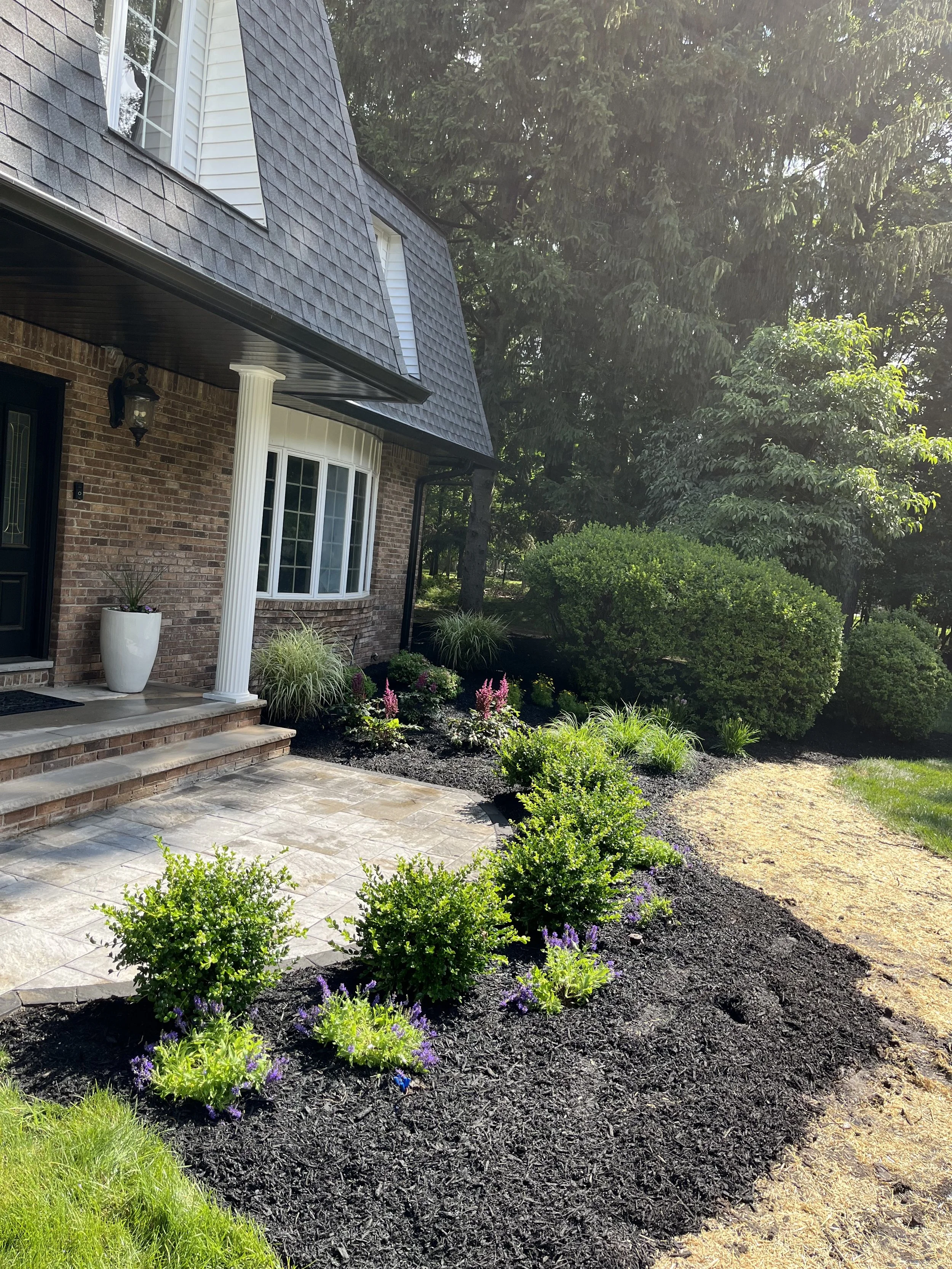 Front yard landscaping of a house, showing a brick porch with stone steps, a black front door, a large white planter with plants, a window with a curved brick border, and a garden bed with various shrubs, flowers, and mulch, with greenery and trees i