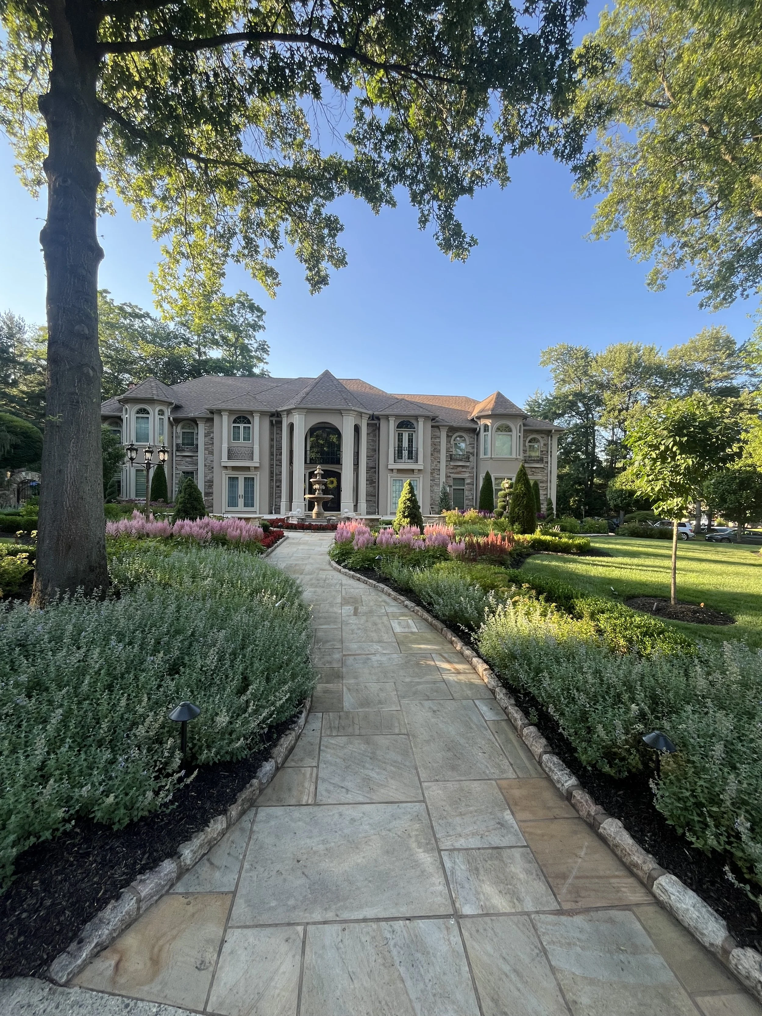 A large, elegant house with a small front yard and a stone walkway leading to its entrance. The house features multiple windows, a fountain in the center of the yard, and is surrounded by lush greenery and flowers. Tall trees frame the view, and a cl