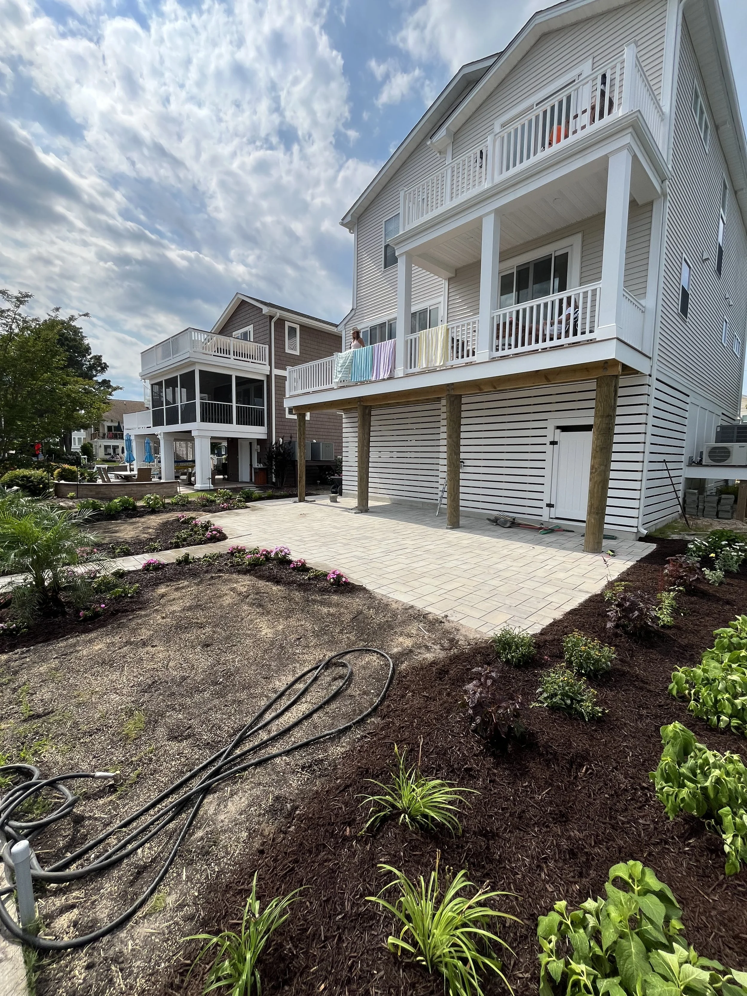 Backyard garden area with newly planted flowers and plants, surrounded by mulched soil, paths, and a hose, with a multi-story house in the background under a partly cloudy sky.