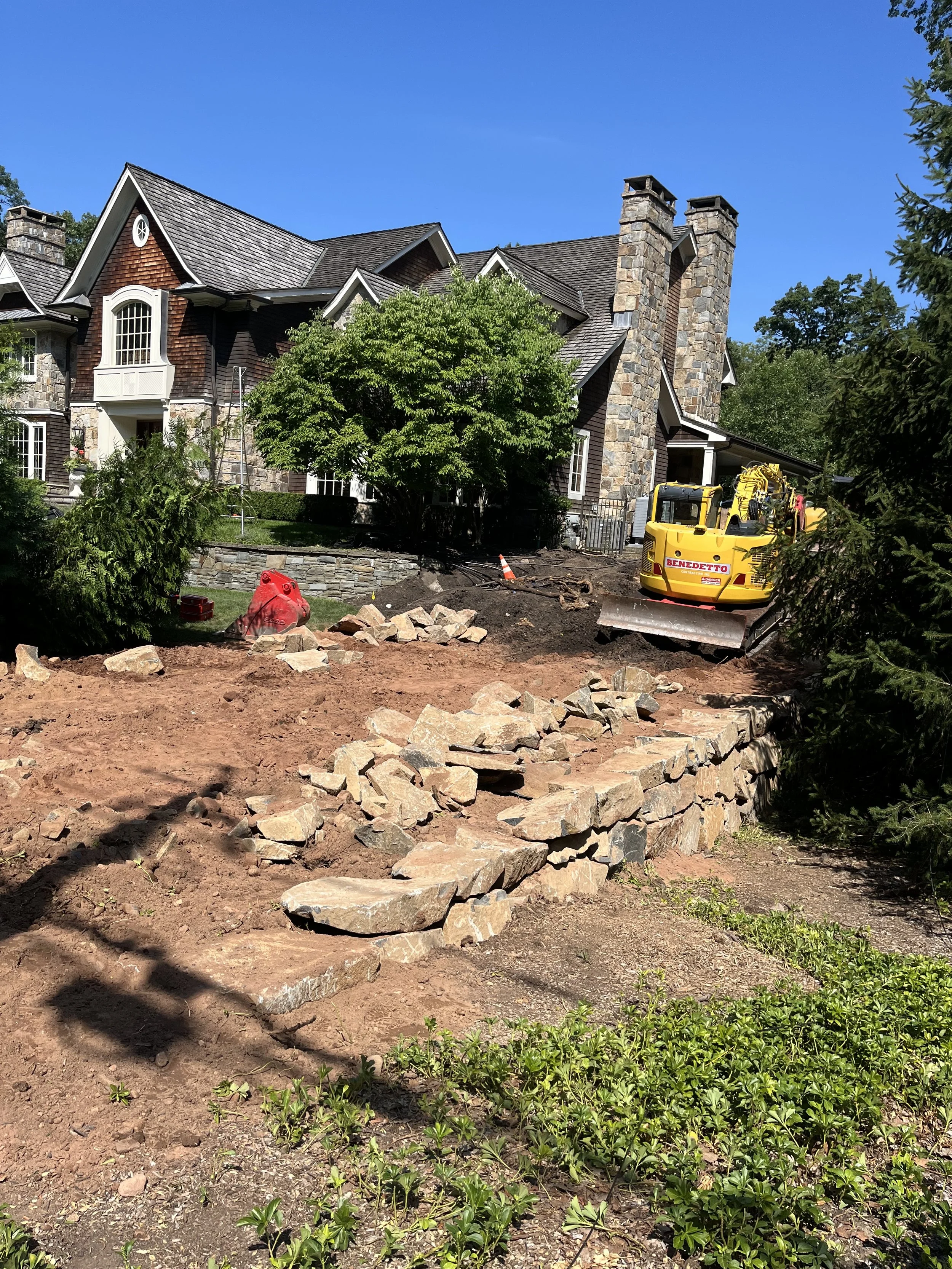 A house under construction with a large yard, a yellow construction vehicle, dirt and rocks, and a tree in front of the house on a sunny day.