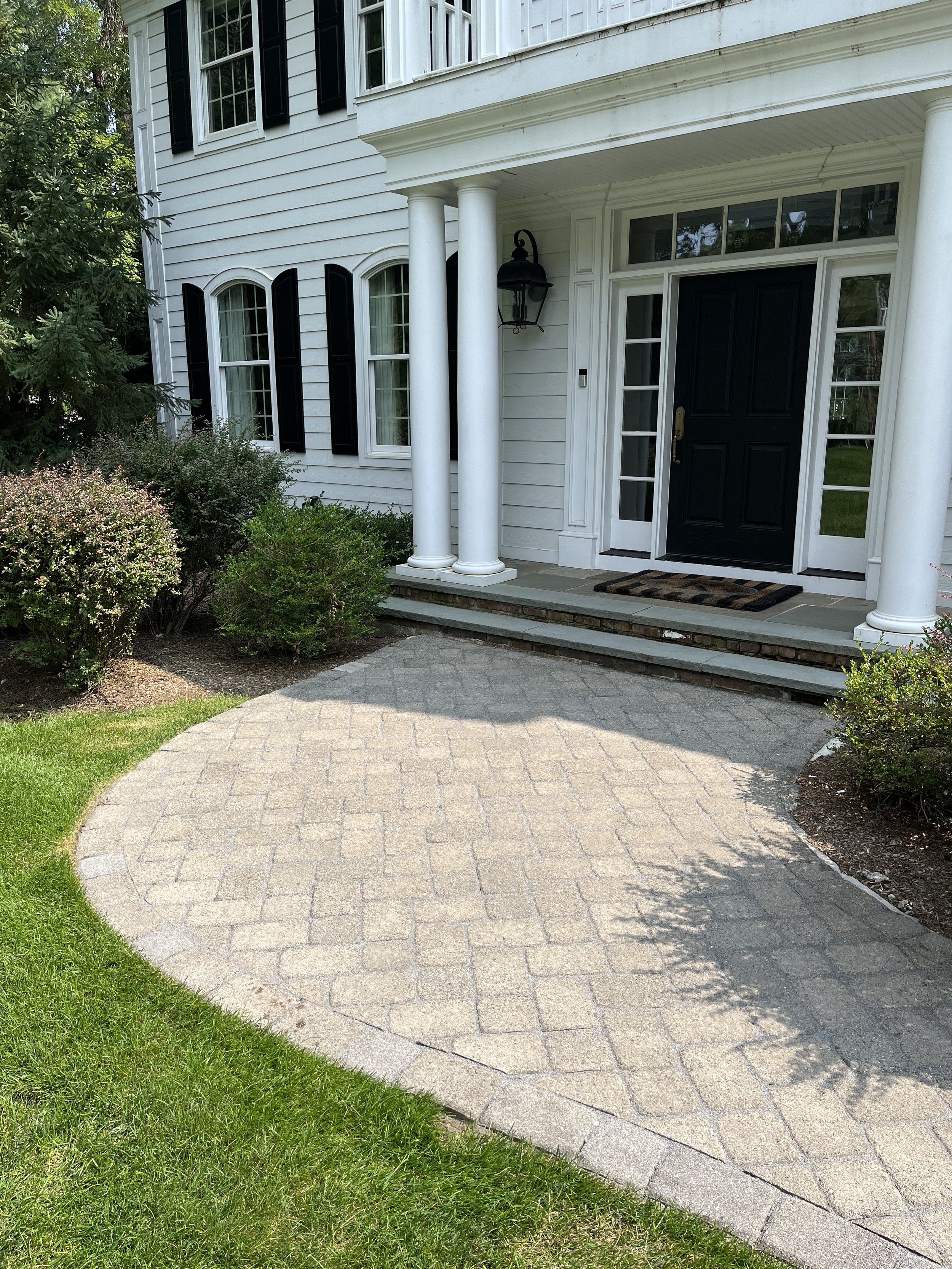 Front porch of a white house with black front door, black shutters, white columns, and a brick step staircase. There are shrubs and a curved brick pathway leading to the porch.