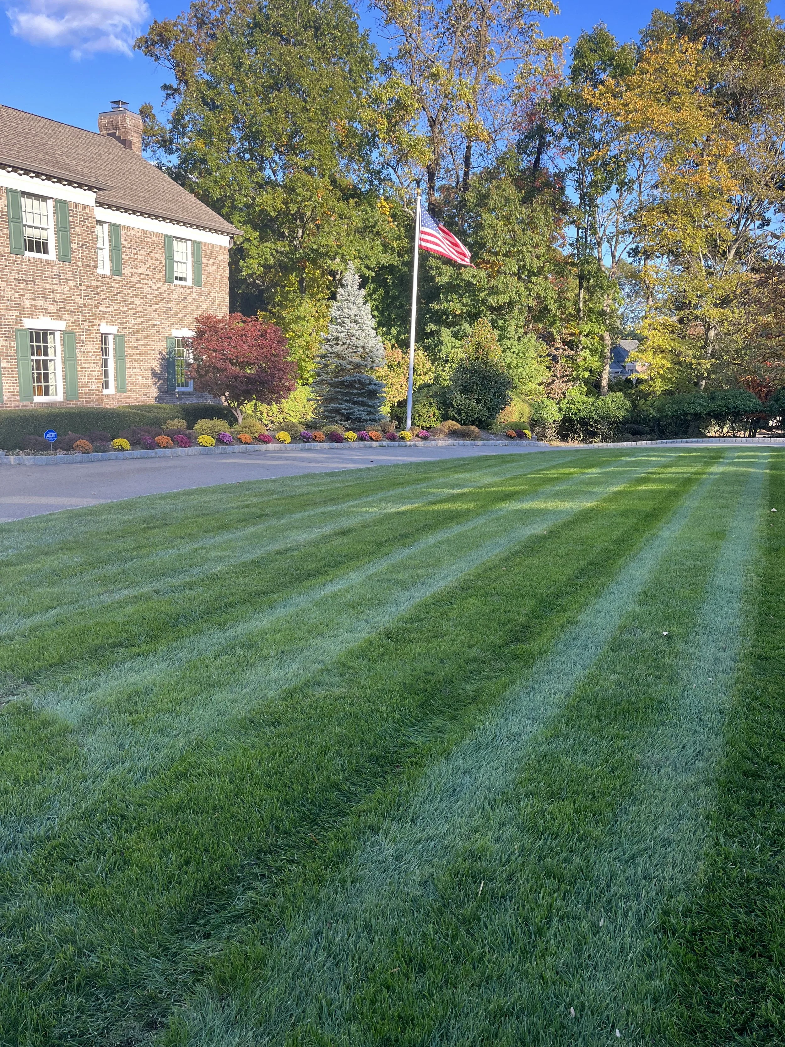 A well-maintained front yard with lush green grass, colorful flower beds, a brick house with green shutters, and a tall American flag on a flagpole. There are trees with autumn foliage in the background under a clear blue sky.
