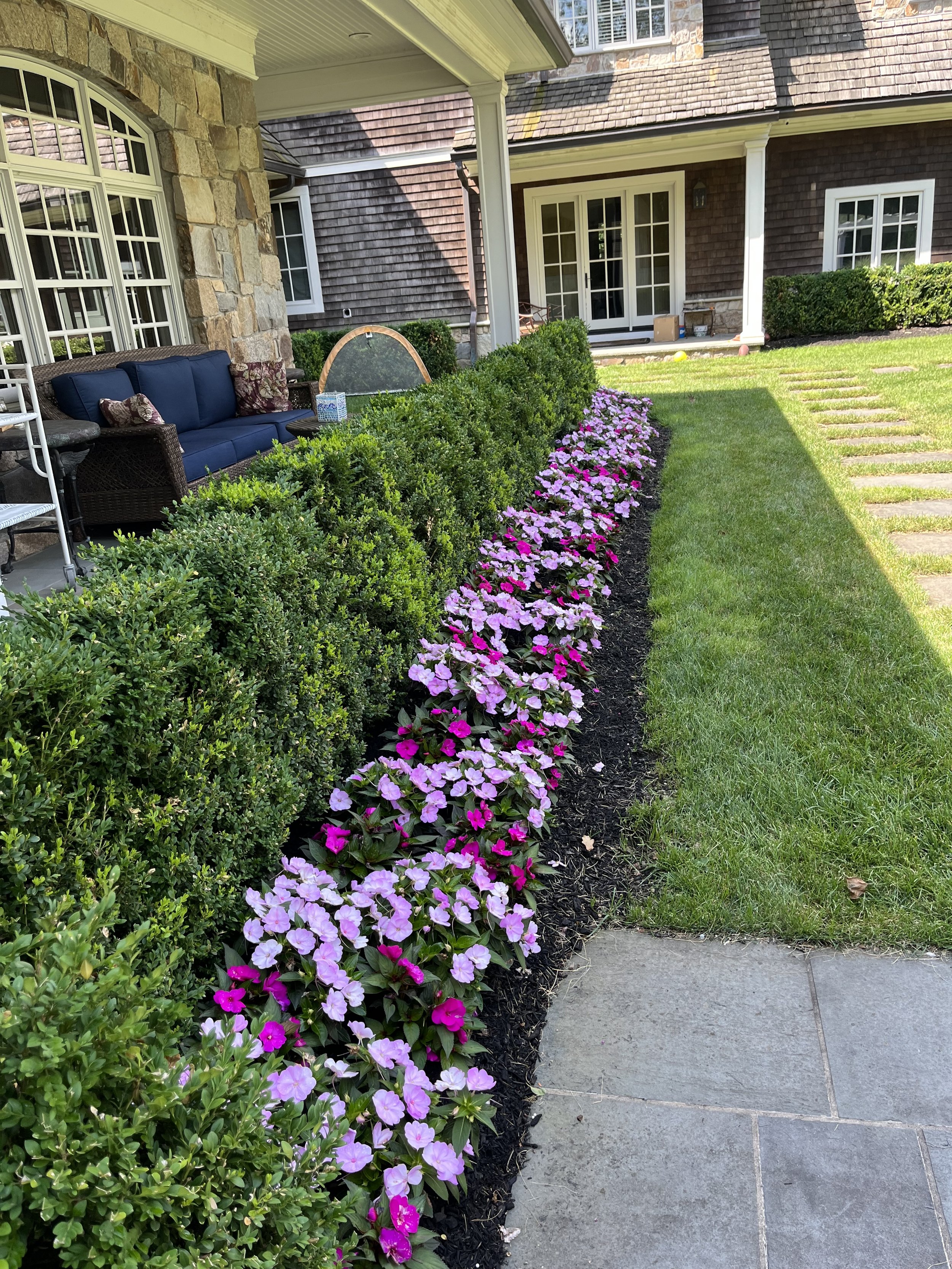 A well-maintained garden with a row of pink and purple flowers lining a pathway next to green bushes. The house's exterior features stone and wooden siding, with large windows and a covered porch area.