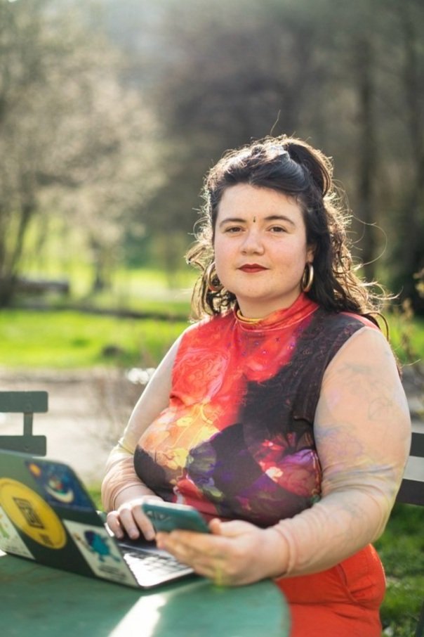 Une femme assise à une table en plein air, tenant un téléphone portable, avec un ordinateur portable devant elle, dans un parc avec des arbres et un chemin en arrière-plan. Agence de communication freelance paris inclusif et engagé