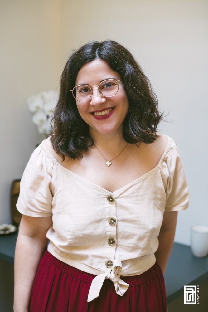 Femme souriante avec lunettes, cheveux ondulés, portant une blouse beige à boutons et une jupe rouge, debout dans une pièce avec une table en arrière-plan. Agence de communication freelance paris inclusif et engagé