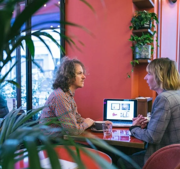 Agence Alan, la communication pour freelance atypiques. Ethique, safe et inclusive. LGBT friendly. Deux freelances discutent dans un café, avec des boissons sur la table.