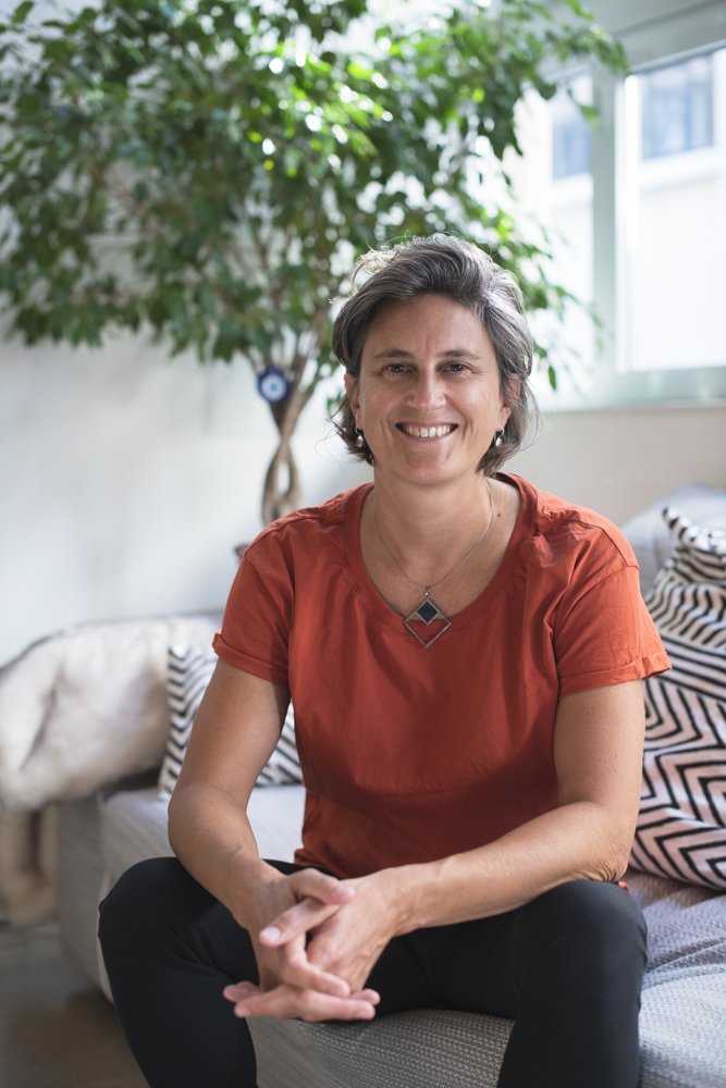 Femme assise sur un canapé en souriant dans un salon lumineux avec un arbre en arrière-plan. Agence de communication freelance paris inclusif et engagé