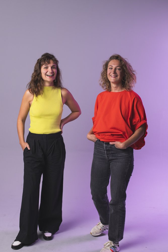Deux femmes posant joyeusement, l'une en haut de couleur jaune, l'autre en haut rouge, avec un fond violet clair. Communication inclusive pour freelance engagé