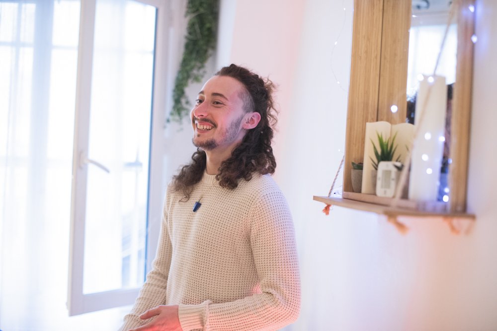 Jeune homme avec long cheveux, souriant dans une pièce lumineuse avec des décorations et plantes. Agence de communication freelance paris