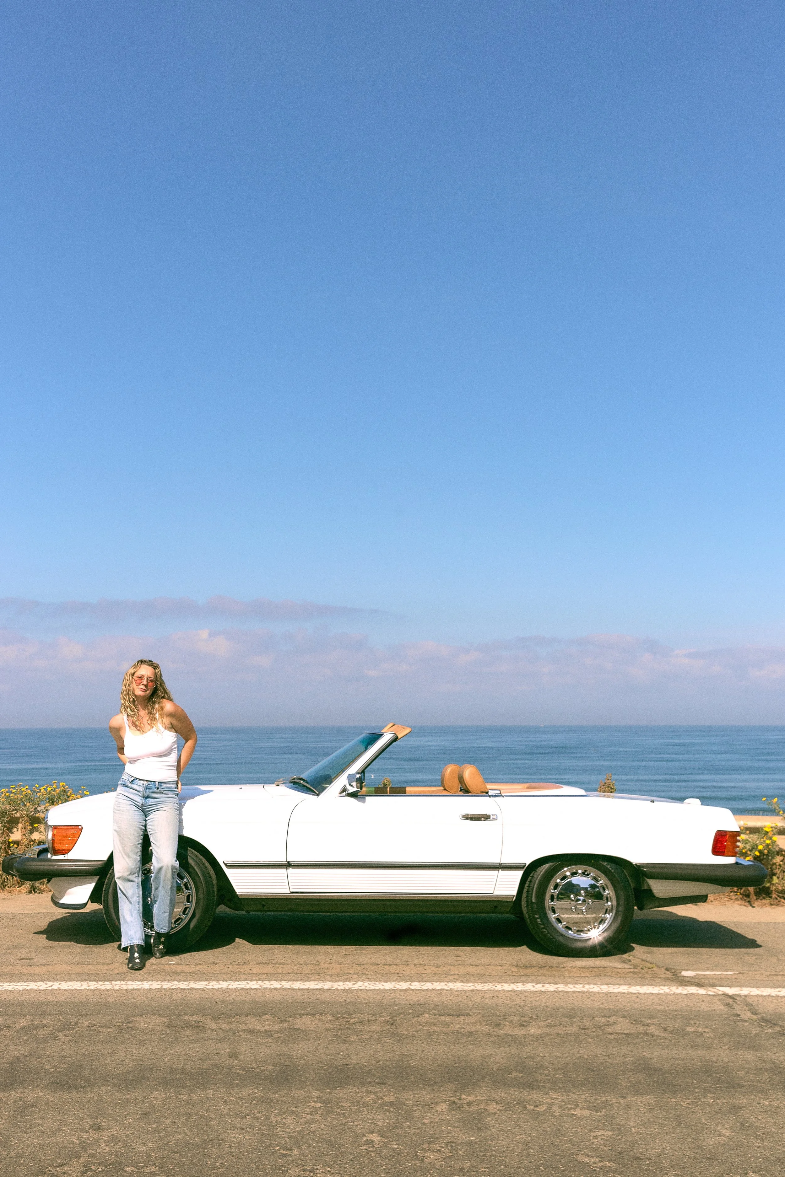 A woman with curly blonde hair, wearing a white tank top, light blue jeans, and black boots, standing next to a white convertible car parked on the side of a road near the beach. The car has a tan interior, and the convertible top is down. The backgr