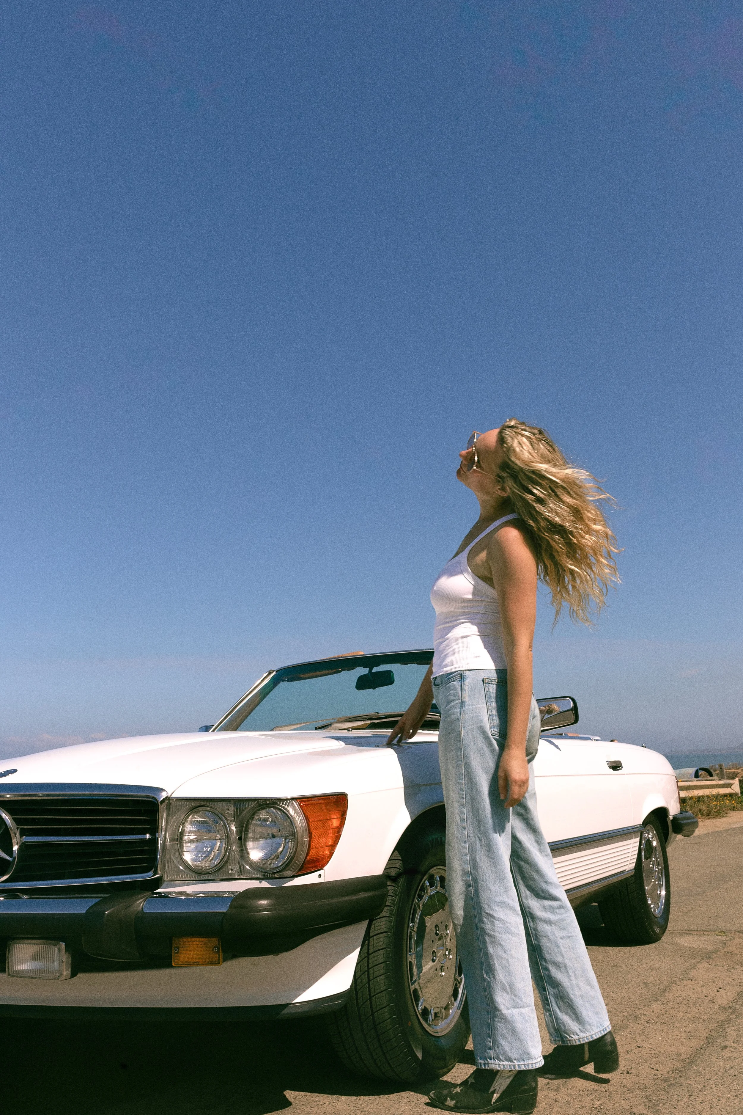 A young woman with long wavy blonde hair wearing sunglasses, a white tank top, and baggy jeans stands beside a white vintage convertible car on a sunny day. The woman appears to be enjoying the outdoor scene with a clear, blue sky overhead.