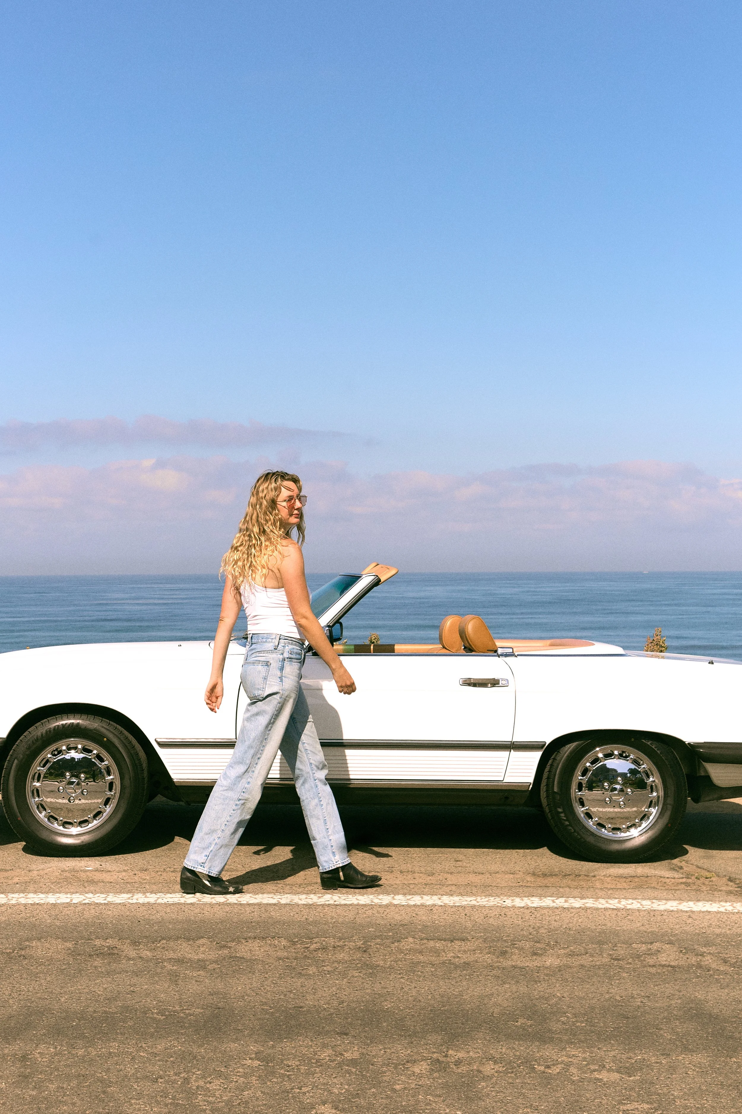 A woman with long curly blonde hair, wearing glasses, a white tank top, and baggy jeans, crosses the street next to a white convertible car while holding a book, with a scenic ocean view and clear blue sky in the background.