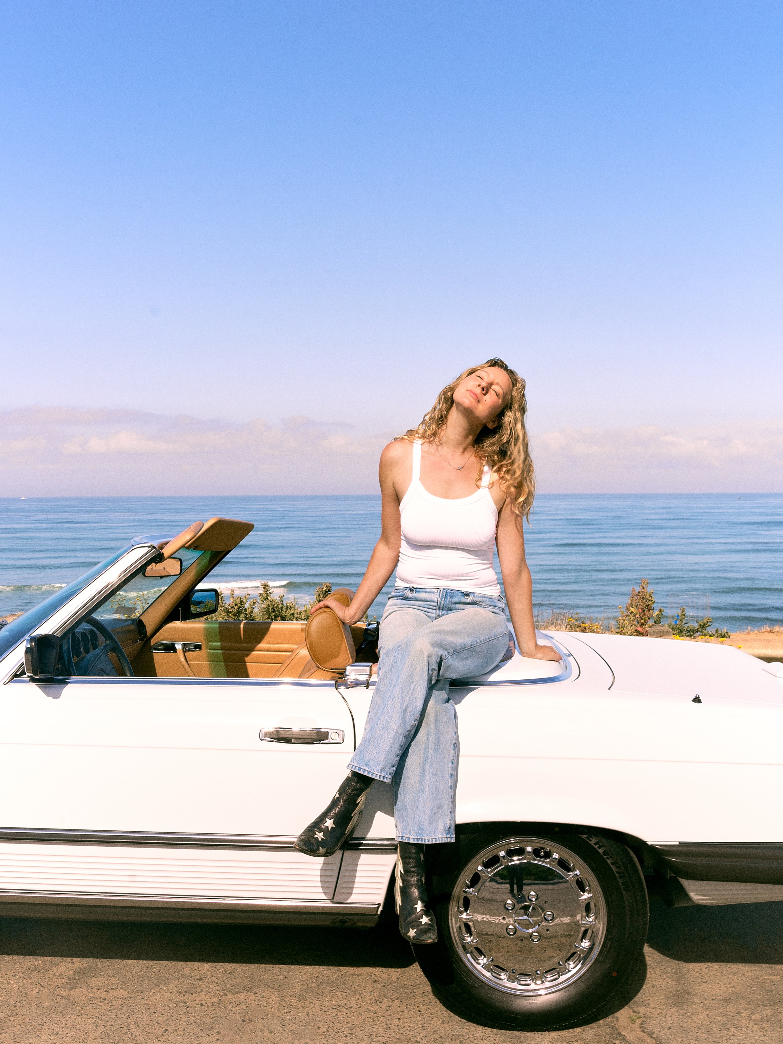 A woman with blonde curly hair, wearing a white tank top, light blue jeans, and black star-patterned boots, sitting on the door of a vintage white convertible car near the ocean under a blue sky.