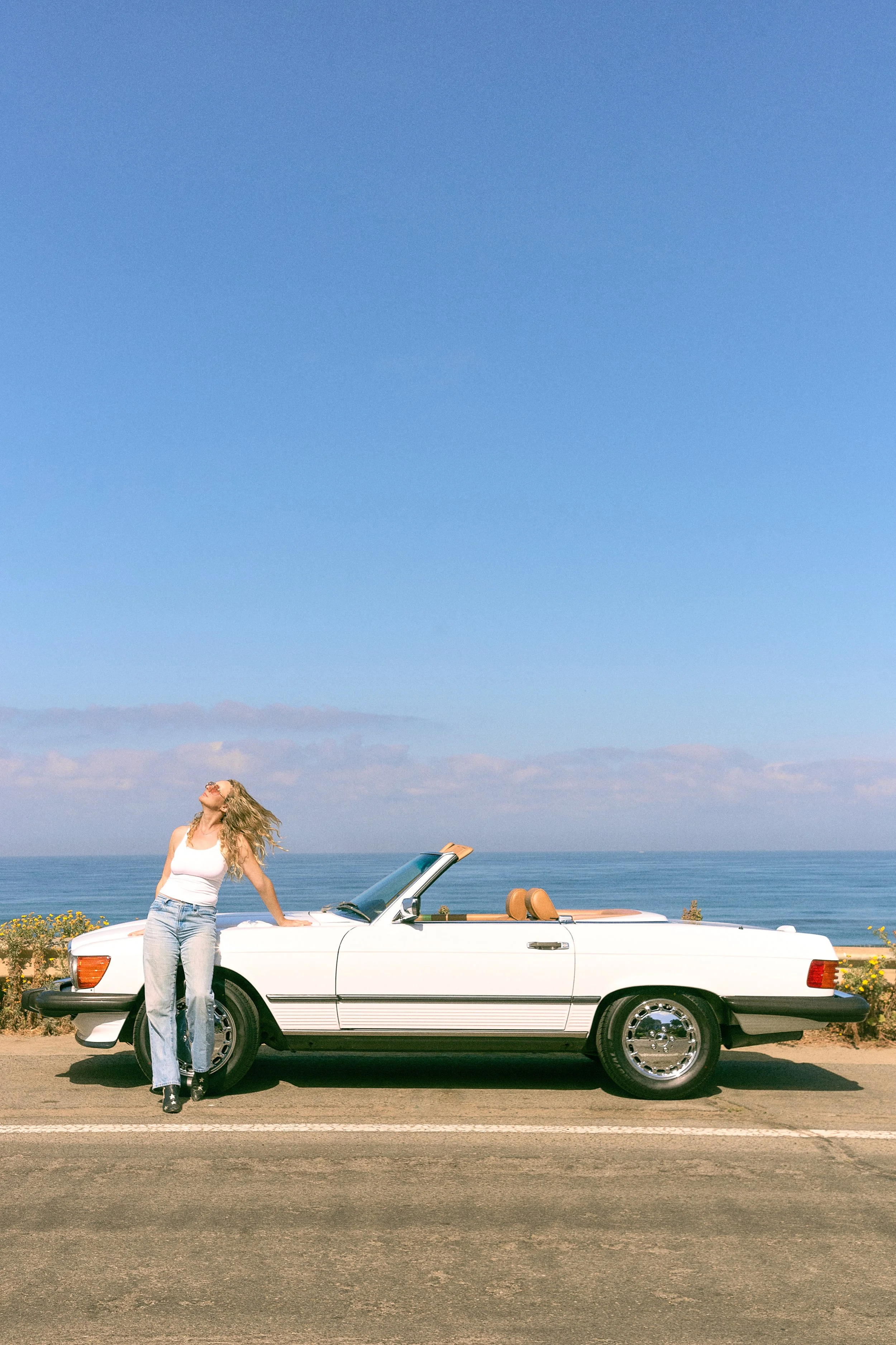 A woman with long blonde hair, wearing a white tank top and jeans, leaning against a white convertible car with the ocean and blue sky in the background.