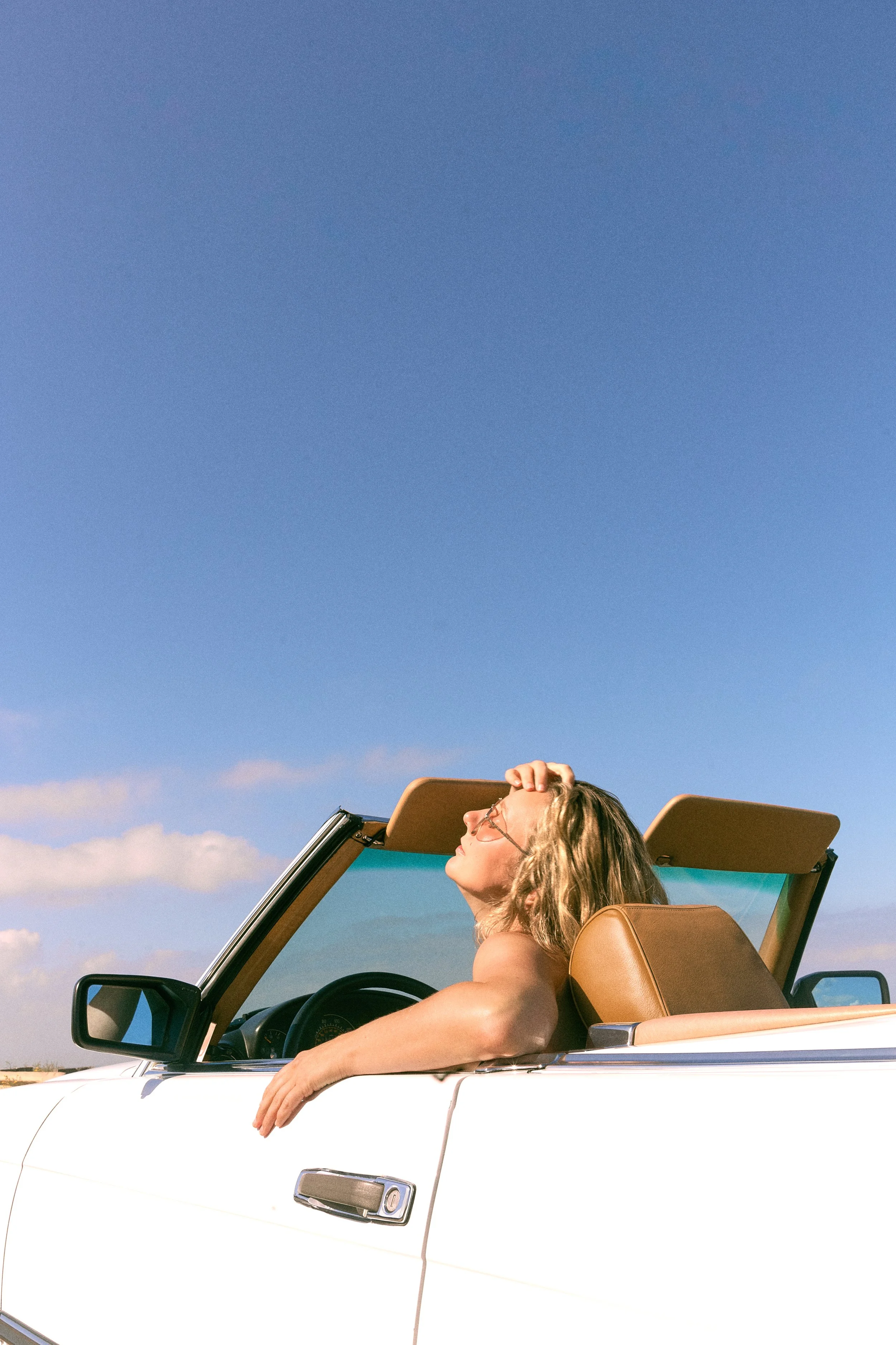 A woman with blonde hair and sunglasses relaxing in a white convertible car with tan interior, sunroof open, under a blue sky with some clouds.