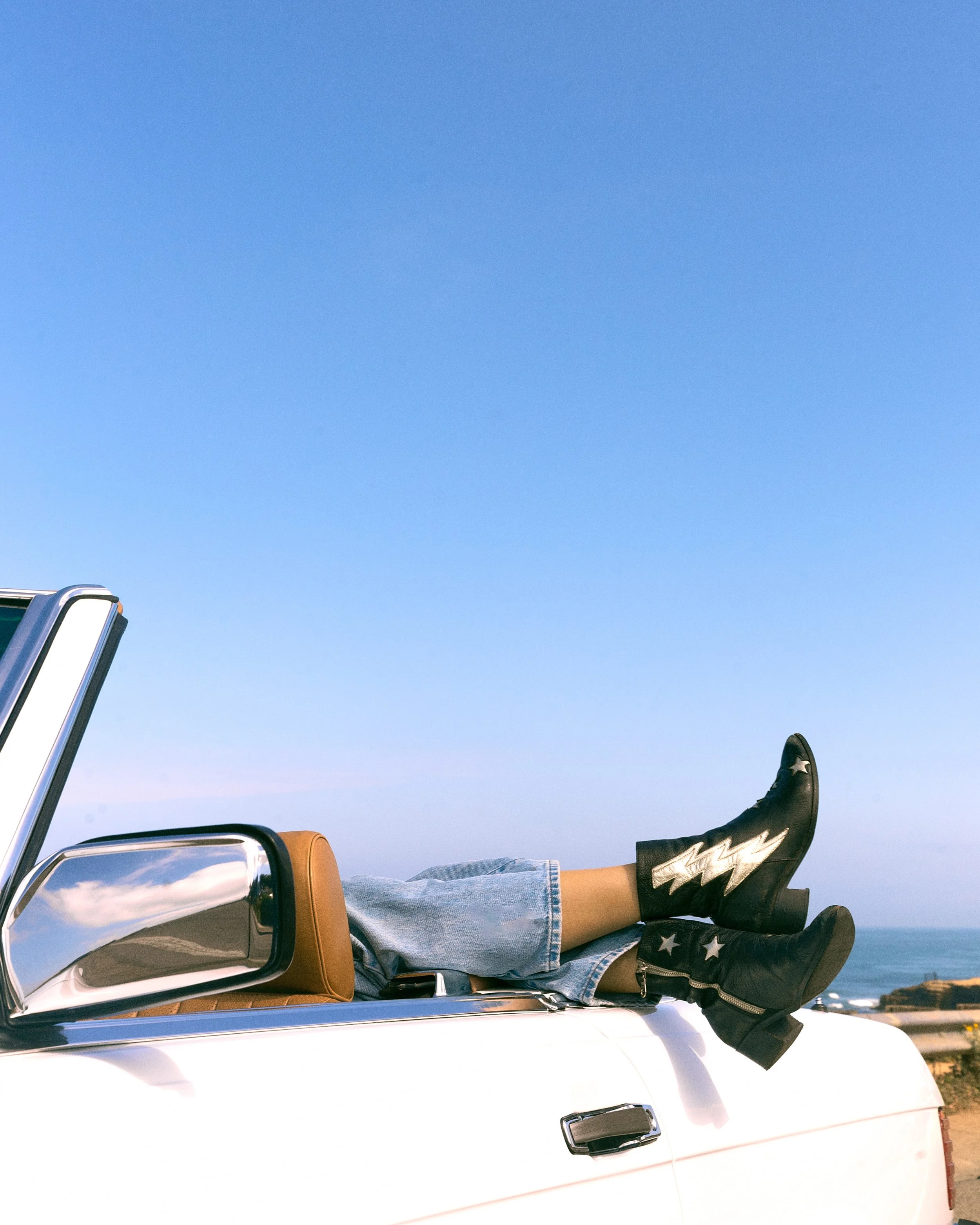 Person lying on the hood of a white car with legs crossed, wearing jeans and black boots with star details, under a clear blue sky.