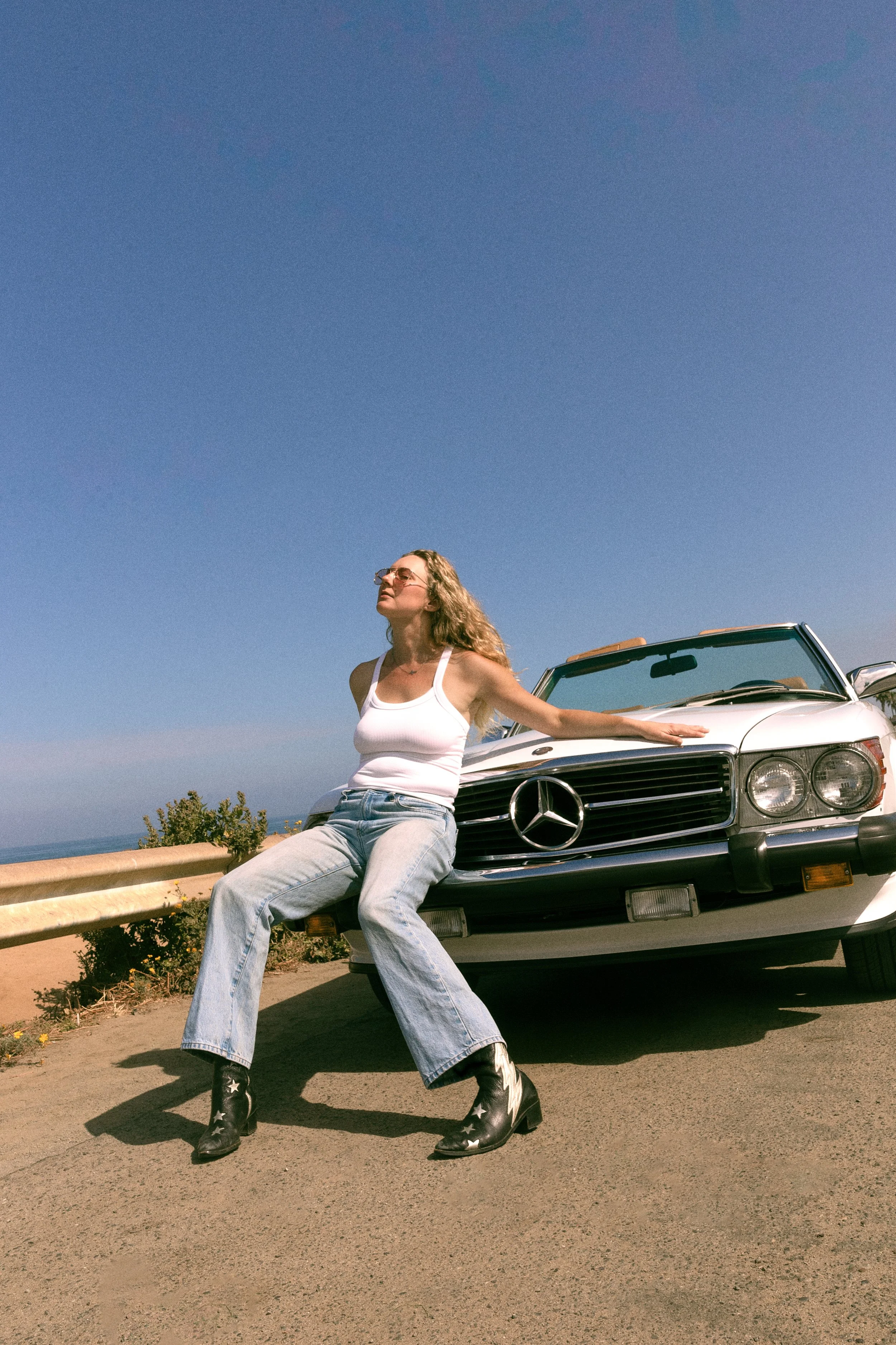 A woman wearing sunglasses, a white tank top, ripped jeans, and star-printed black boots sitting on the front bumper of a vintage Mercedes-Benz convertible car parked on a roadside, with a clear blue sky above and some greenery in the background.