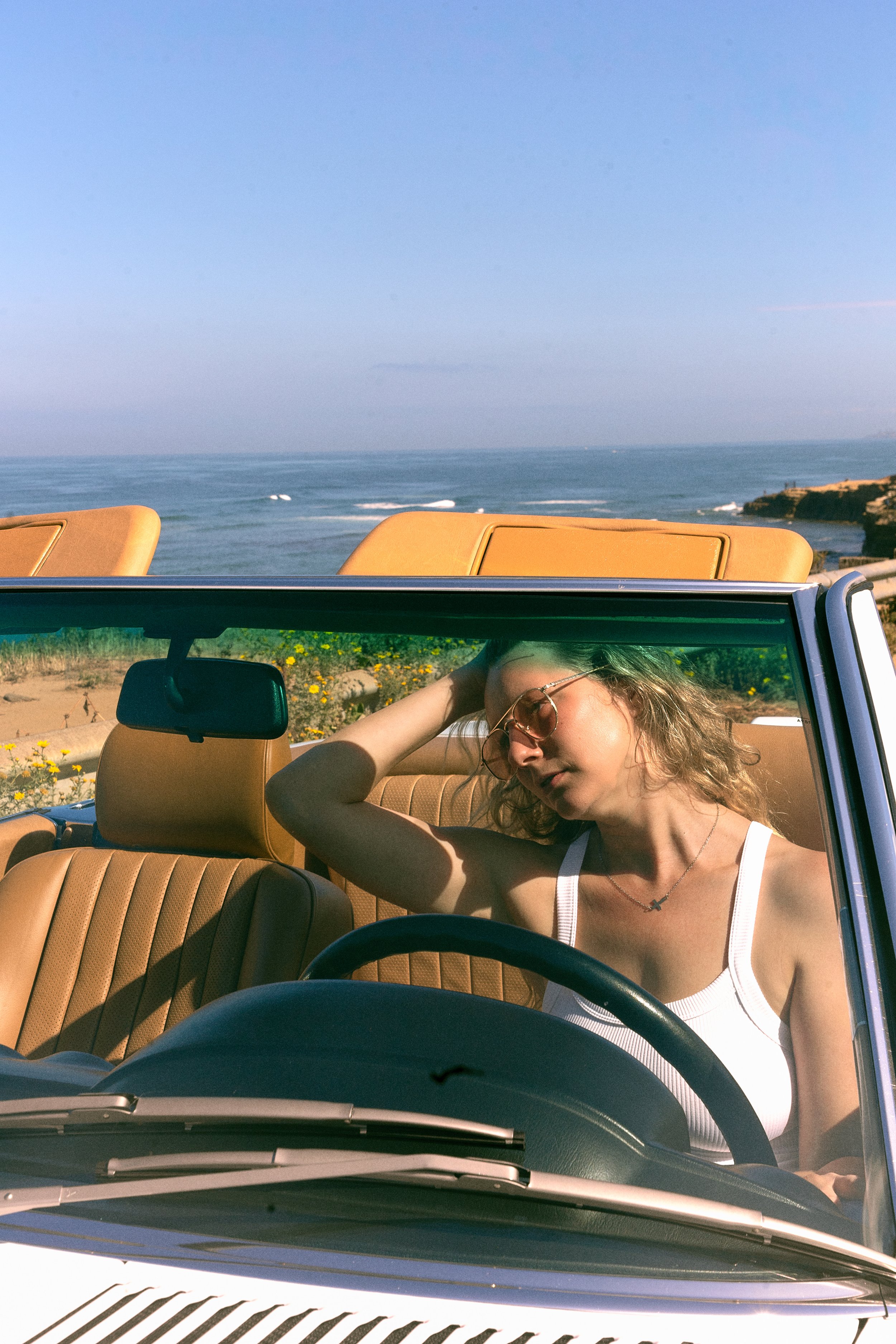 A woman with curly hair and sunglasses relaxing in a tan convertible car parked near a coastline with the ocean and a rocky outcrop in the background.