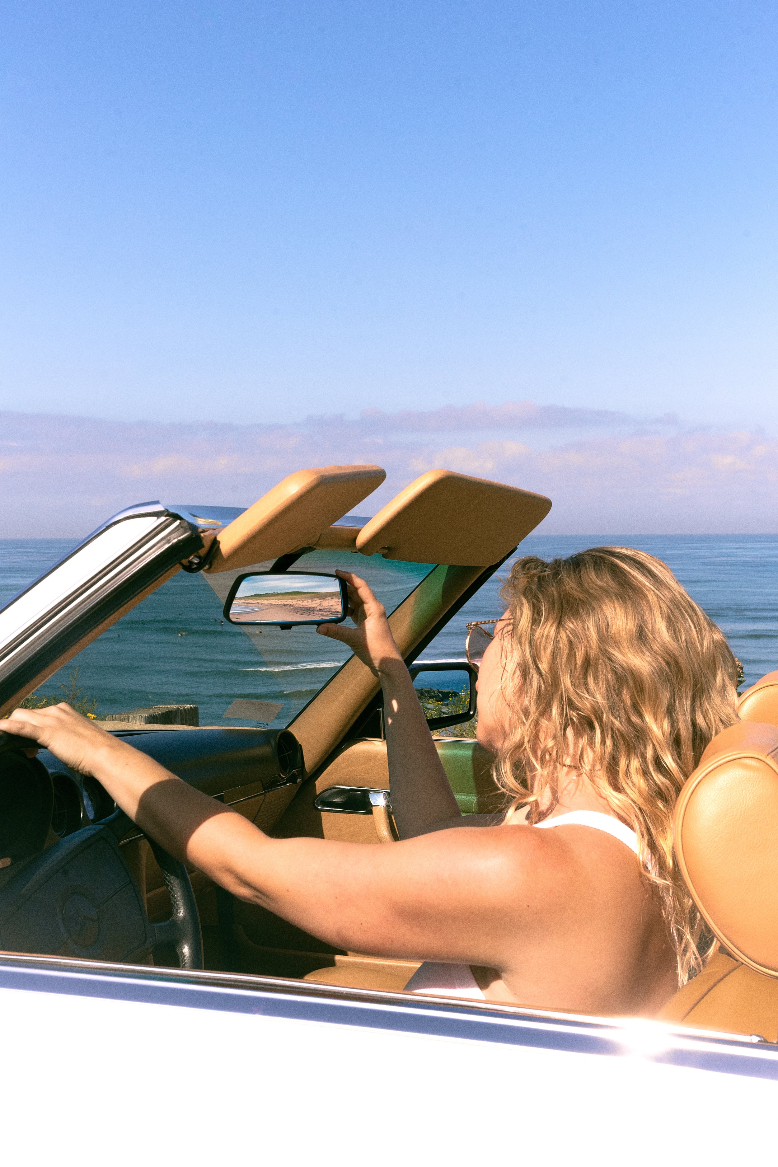 A woman with curly blonde hair, wearing glasses and a white top, is sitting in the driver's seat of a convertible car with tan interior. She is taking a photo with her right hand of the ocean and coastline visible through the windshield and side wind