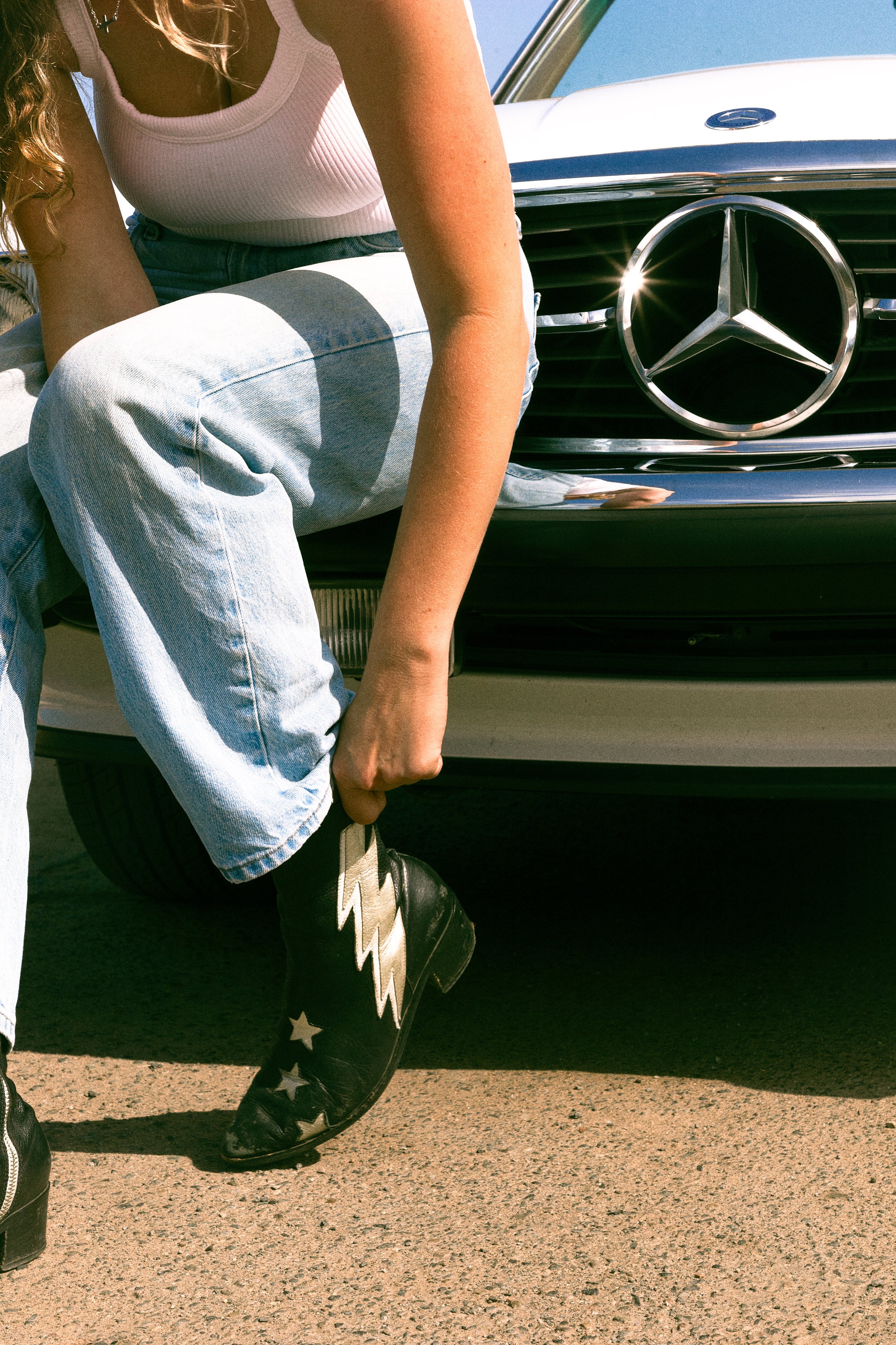 A person kneeling beside a white Mercedes-Benz car with a prominent silver Mercedes emblem on the front grille. The person is wearing a white tank top, light-washed jeans, and black cowboy boots decorated with white stars and a lightning bolt pattern