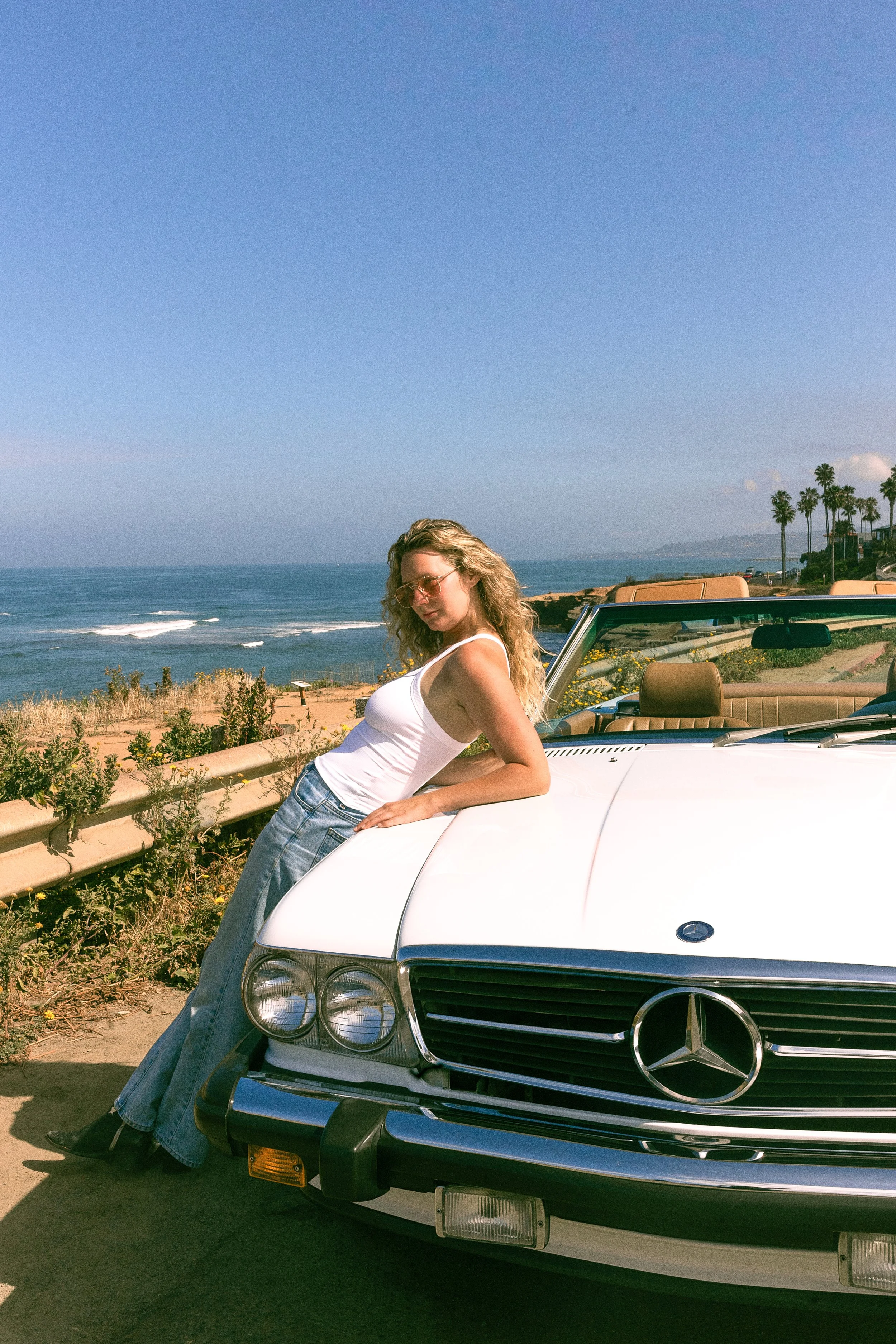 A woman with long blonde hair and sunglasses leaning against a white Mercedes-Benz convertible car with the ocean in the background on a sunny day.