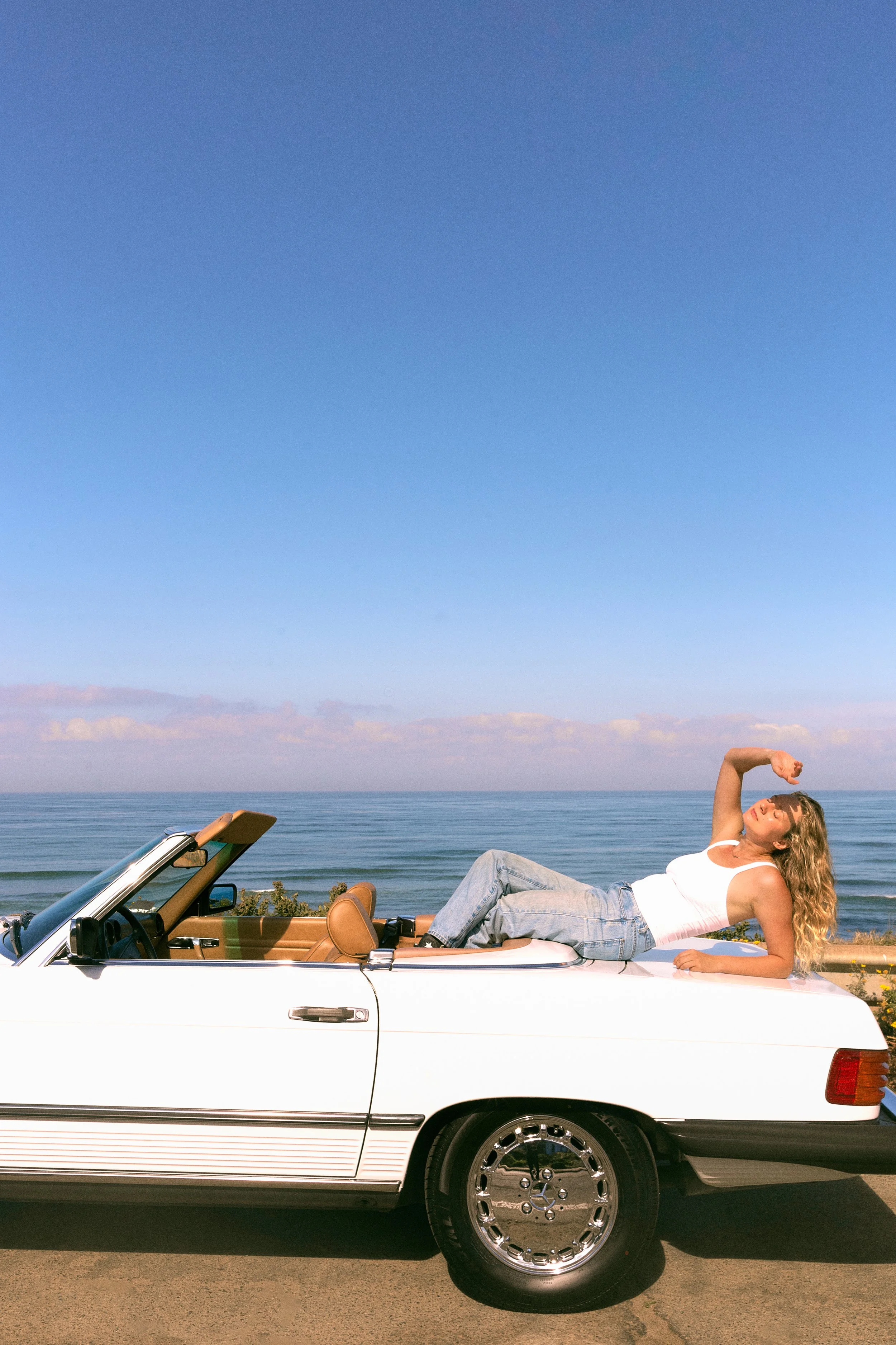 A woman with long wavy blonde hair lying on the back seat of a white convertible car parked by the ocean, holding her right arm up in a relaxed pose, wearing a white tank top and blue jeans, with the ocean and sky in the background.