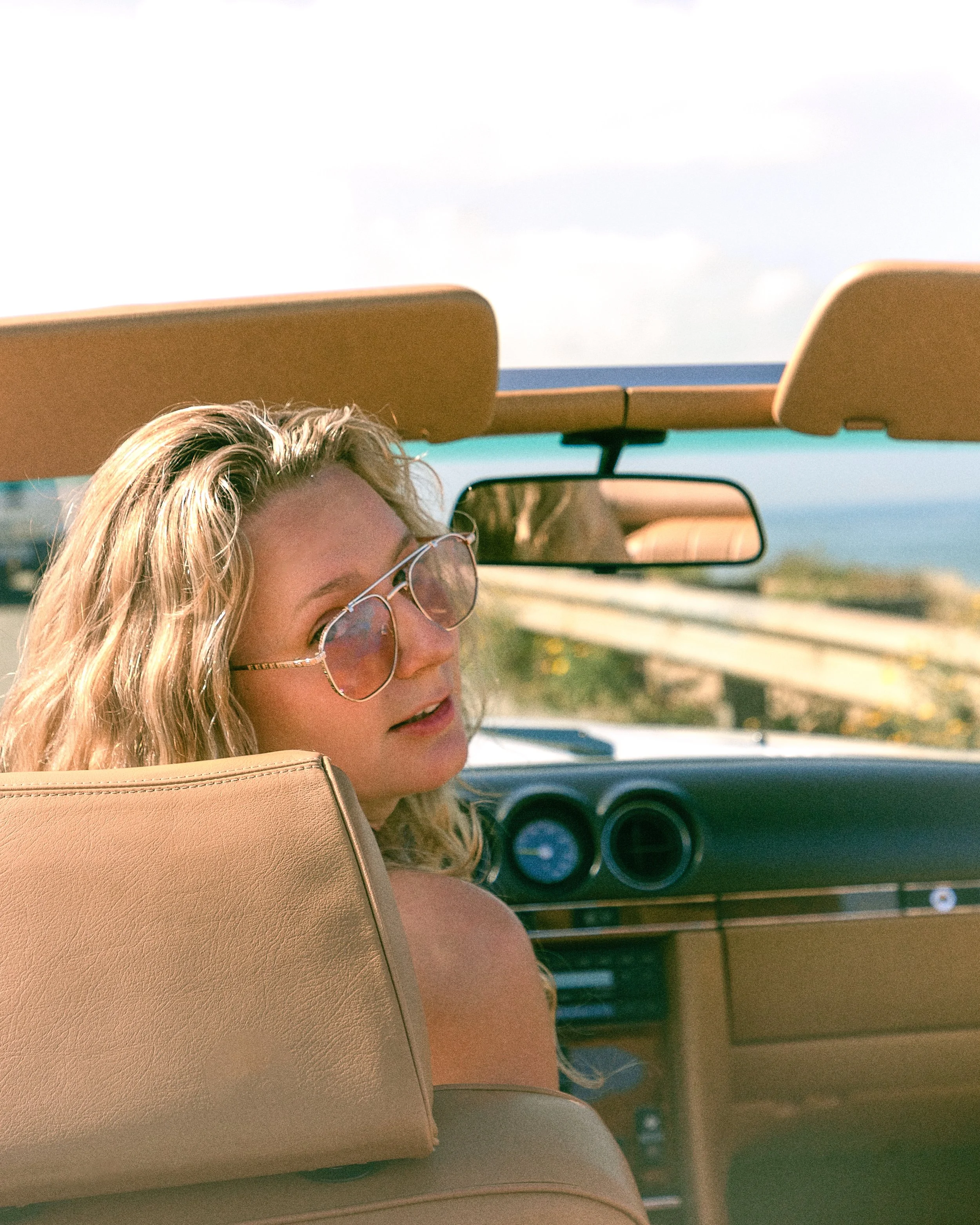 A woman with blonde hair and sunglasses riding in the backseat of a convertible car, with a rearview mirror visible, driving along a highway with a guardrail and a blue sky.