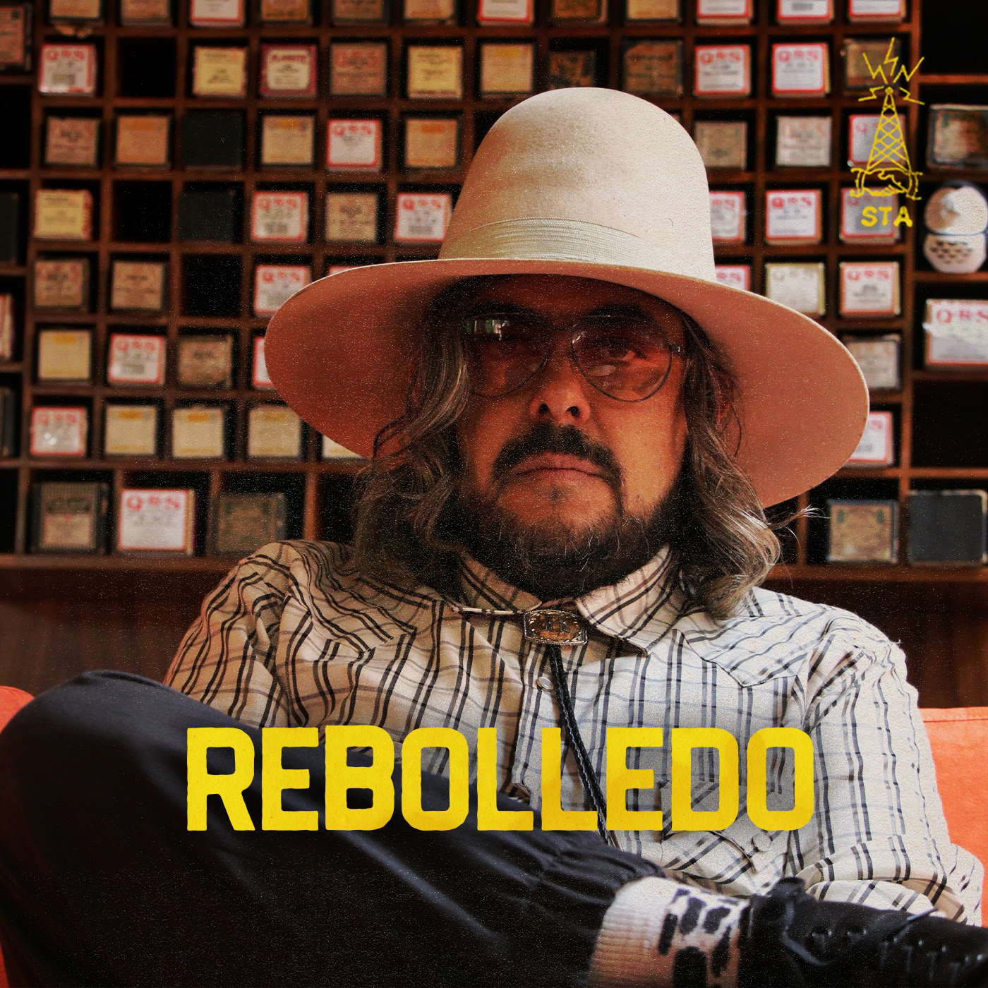 A man (dj)  with long hair, glasses, and a beard wearing a wide-brimmed beige hat and a plaid shirt, sitting in front of a background of small boxes with labels. The word 'REBOLEDO' is written in bold yellow letters at the bottom of the image.