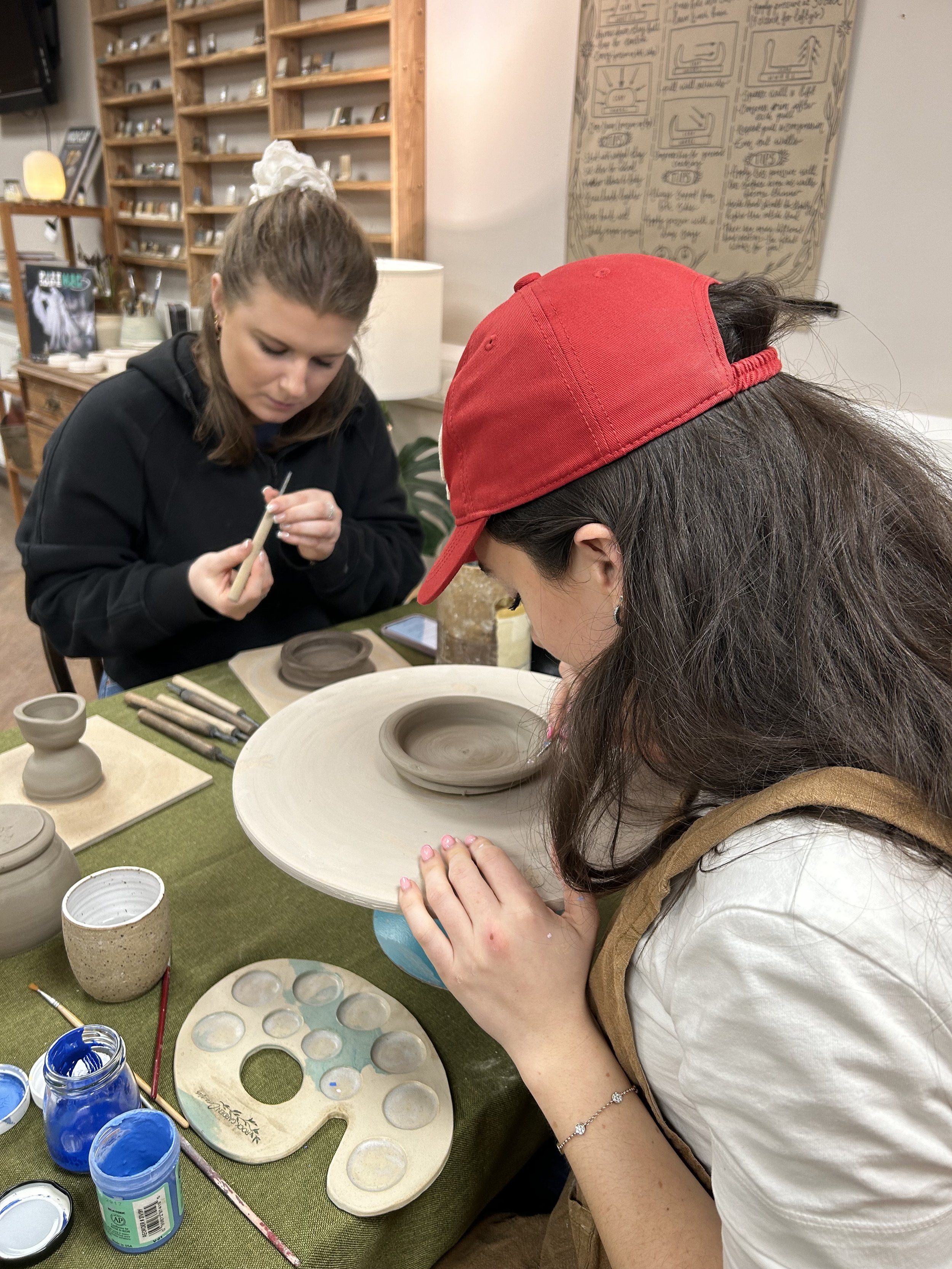 Two women are working on a pottery wheel, shaping a clay bowl, with various pottery tools and paint at the table.