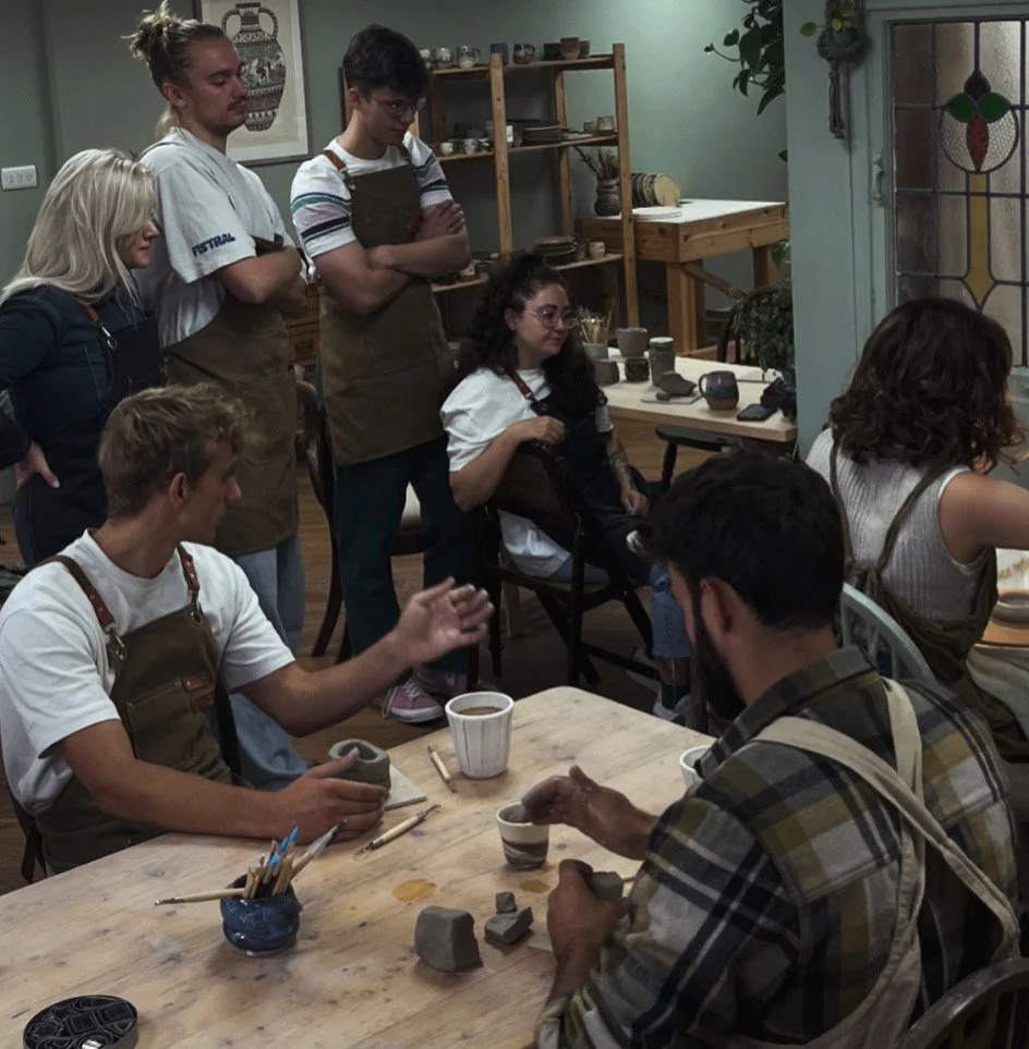 People gathered in a pottery studio, working on clay projects and observing others shaping clay.