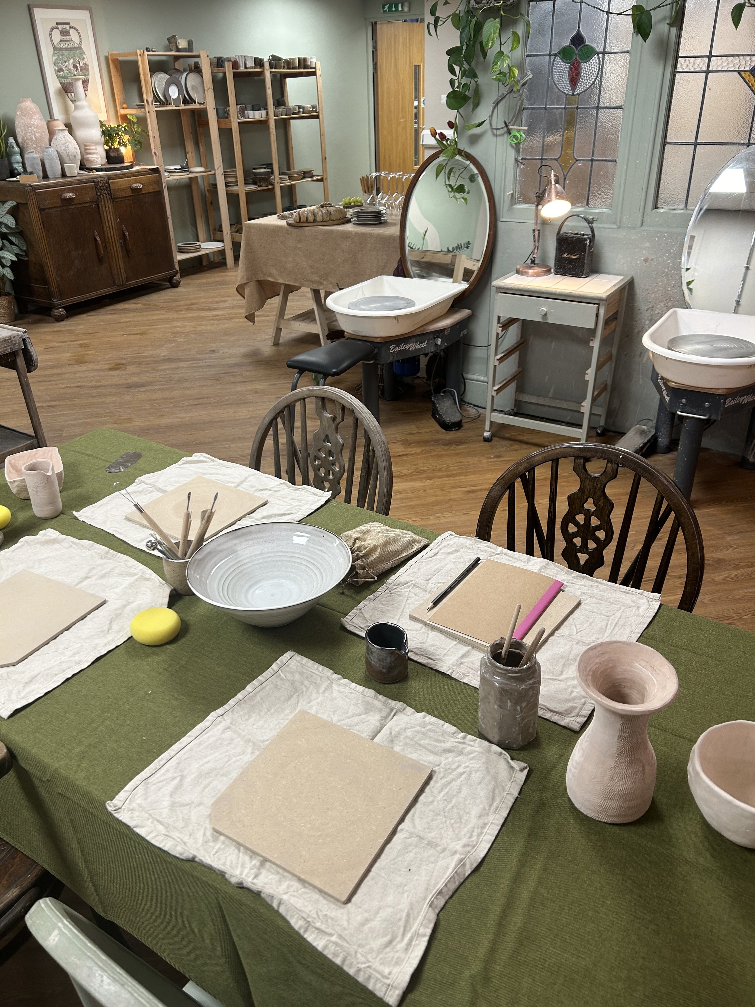 A pottery studio with a table set for a class, featuring plates, bowls, and pottery tools, and pottery wheels in the background near a window.