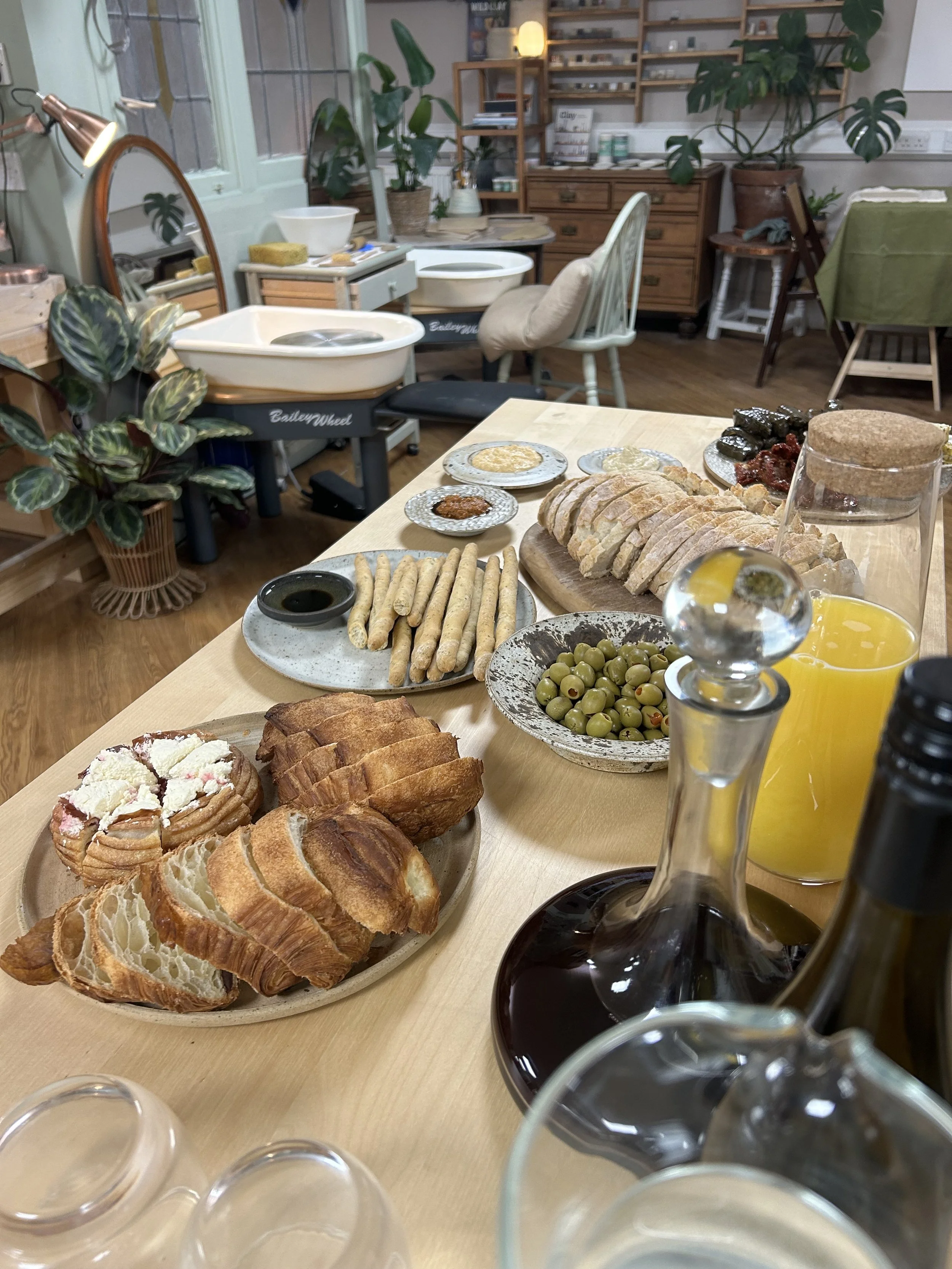 A table with assorted breakfast foods including sliced bread, pastries, green olives, and juice in a cozy, well-lit room with plants and furniture in the background.
